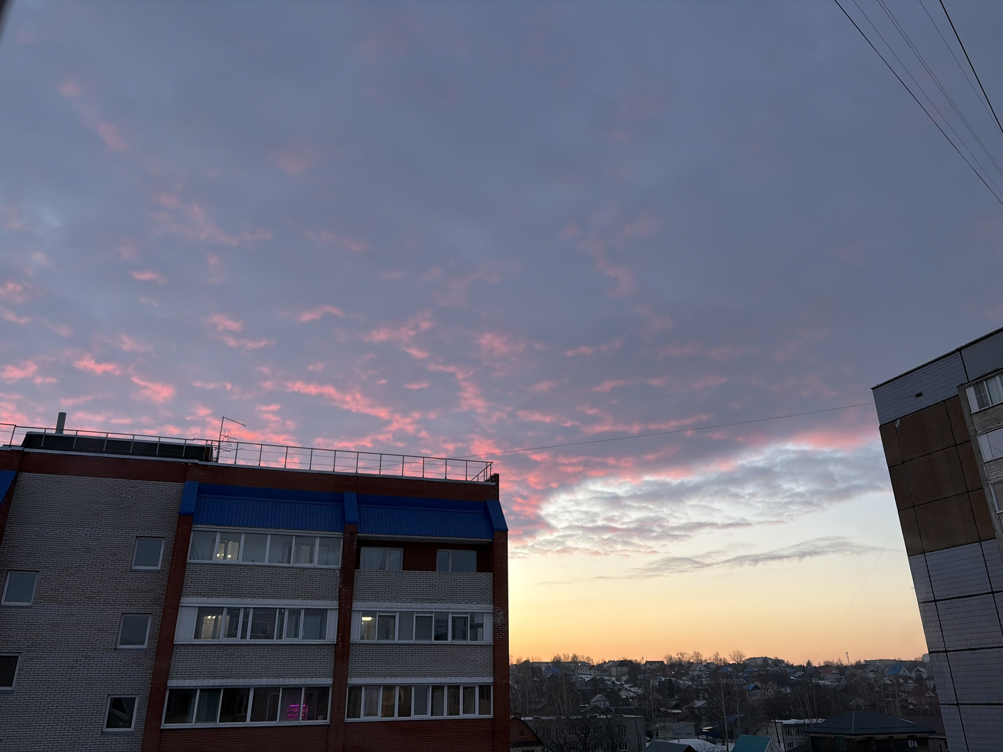 The image captures an urban landscape in Izhevsk, Russia, at either sunrise or sunset. The sky dominates the upper two-thirds of the frame, displaying a gradient from deep blue-grey at the zenith to an orange-yellow hue near the horizon. Numerous small, scattered altocumulus clouds are illuminated with shades of pink and red, particularly in the upper-left quadrant.
In the foreground, a multi-story building, likely residential, occupies the left side of the frame. Its facade consists of reddish-brown brickwork on the left portion and lighter grey/white panels on the right, interspersed with vertical red brick columns. The top floor features a prominent blue sloped roof or architectural trim, and a metal railing runs along the roofline. A dark, vertical pipe or vent extends from the rooftop. Multiple windows are visible, some reflecting light, while interior lights are active in several lower windows, one appearing magenta.
A section of a second multi-story building, constructed with large, light-grey and dark-grey rectangular panels, is visible on the far right edge of the frame.
Between the two buildings, a sprawling residential area extends to the horizon. This area features numerous detached houses with varying roof colors, including blue, grey, and white. Bare trees, suggesting a winter or late autumn season, are interspersed throughout the houses. Patches of light-colored ground, indicative of snow, are visible among the buildings and trees.
Several dark, thin utility lines or power cables traverse the upper-right portion of the sky, running diagonally downwards from the top-right corner. There are no people visible in the scene.