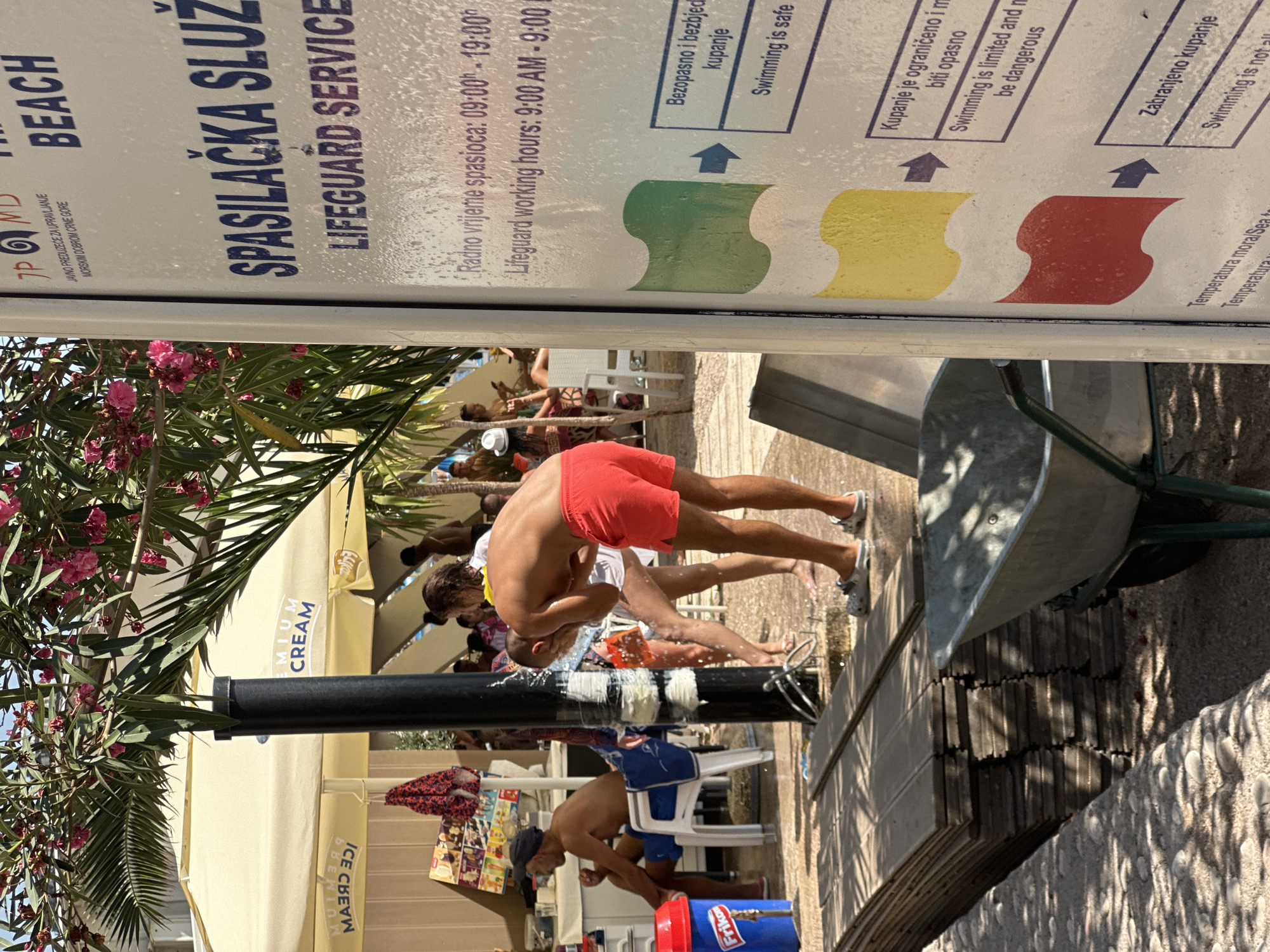 An outdoor scene in Budva, Montenegro, features a lifeguard service sign with multilingual information, including Croatian and English. The sign displays different colored flags and their corresponding safety advisories for swimming. To the left of the sign, a man in red shorts is showering at an outdoor showerhead, with water spraying around him. In the background, people are seen relaxing under a large yellow umbrella that reads "CREAM." Further back, other beachgoers and structures are visible. A stack of wooden planks and a wheelbarrow are in the foreground. The ground is covered in gravel and pebbles. Lush green foliage and pink flowers hang overhead, suggesting a warm, sunny day at a beach or resort.