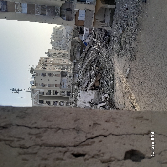 The image captures the aftermath of destruction. Buildings stand damaged with visible cracks and rubble. Concrete debris fills the foreground, leading to a sandy path. The horizon reveals more battered buildings under a pale sky. The scene conveys desolation and infrastructural ruin.