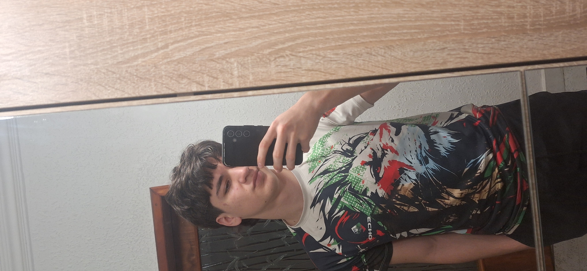 A young man is taking a selfie in a bathroom mirror. The mirror is framed with a light brown wood-grain material, with a white wall in the background. He is wearing a white t-shirt with a large print that includes red, green, and black designs. He is holding a black smartphone with three cameras in his right hand. A dark wooden cabinet is partially visible behind him. The location appears to be indoors, possibly a private residence. There is no indication of Burgas, Bulgaria.