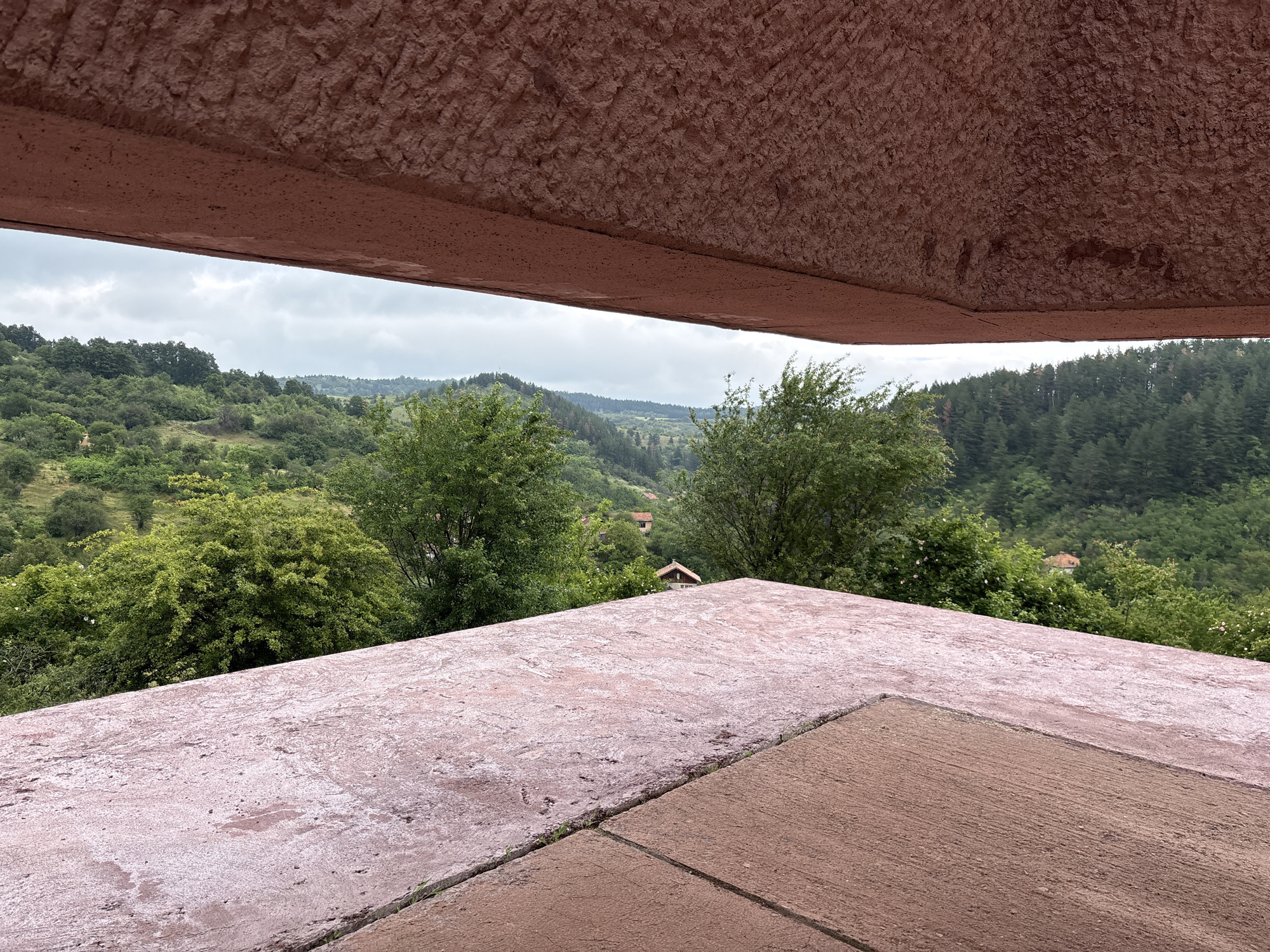 The image depicts a panoramic view of the Gurgulyat, Bulgaria landscape, framed by two prominent, reddish-brown concrete structures. The foreground is dominated by a textured, pinkish-red concrete surface that angles diagonally across the frame, suggesting a rooftop or elevated platform. A darker, reddish-brown concrete surface is visible in the lower right corner, also angled and contributing to the structural elements framing the view.
Beyond these structures, a verdant, rolling landscape unfolds under a cloudy sky. Lush green trees and vegetation cover the hills, with denser forests on the distant slopes. Several small, red-roofed houses are nestled among the trees, indicating a rural or semi-rural setting. The overall impression is one of a natural environment with scattered human habitation, viewed from a man-made vantage point. There are no people or animals visible in the image.