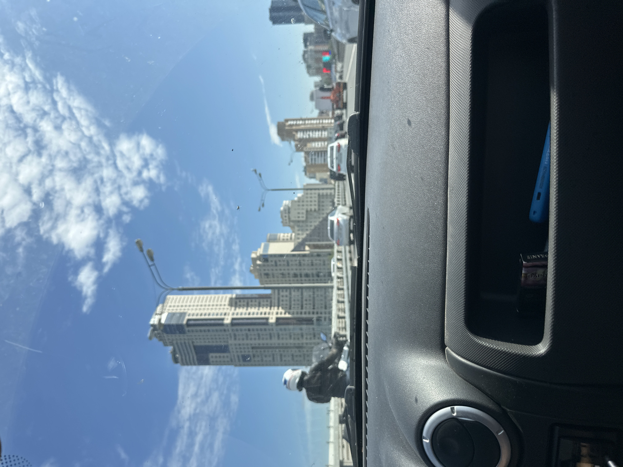 The image, taken from the interior of a vehicle, shows a view through the windshield onto a bright, sunny day in Khimki, Russia. The foreground is dominated by the dashboard of the car, with a storage compartment visible containing a blue object and a brown item that appears to be a pack of cigarettes.
Beyond the car, a busy highway stretches into the distance, filled with various vehicles including cars and a motorcycle. A person wearing a white helmet and a dark jacket is riding the motorcycle in the left lane. Traffic appears to be moving, though some congestion is visible.
The cityscape of Khimki is characterized by numerous tall buildings of varying architectural styles, including a prominent, light-colored skyscraper with a distinctive stepped top. Streetlights and power lines are visible alongside the road. The sky is a clear blue with scattered white clouds. The overall impression is a typical urban scene during daylight hours in Russia.