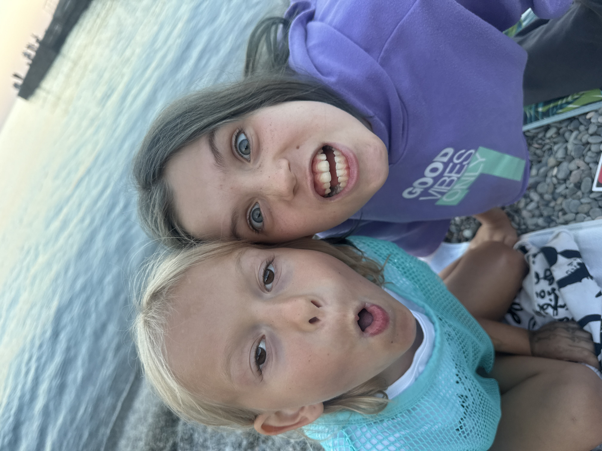 Two young girls are seated on a pebble beach near the water. The girl on the left has blonde hair and is wearing a turquoise mesh top over a white shirt. Her mouth is open in a surprised expression. The girl on the right has greyish-brown hair and is wearing a purple hoodie with the text "GOOD VIBES ONLY" visible. Her eyes are wide and her mouth is open, showing her teeth in a playful grimace. In the background, the sea is visible with a pier in the distance. The overall setting suggests an outdoor recreational area.