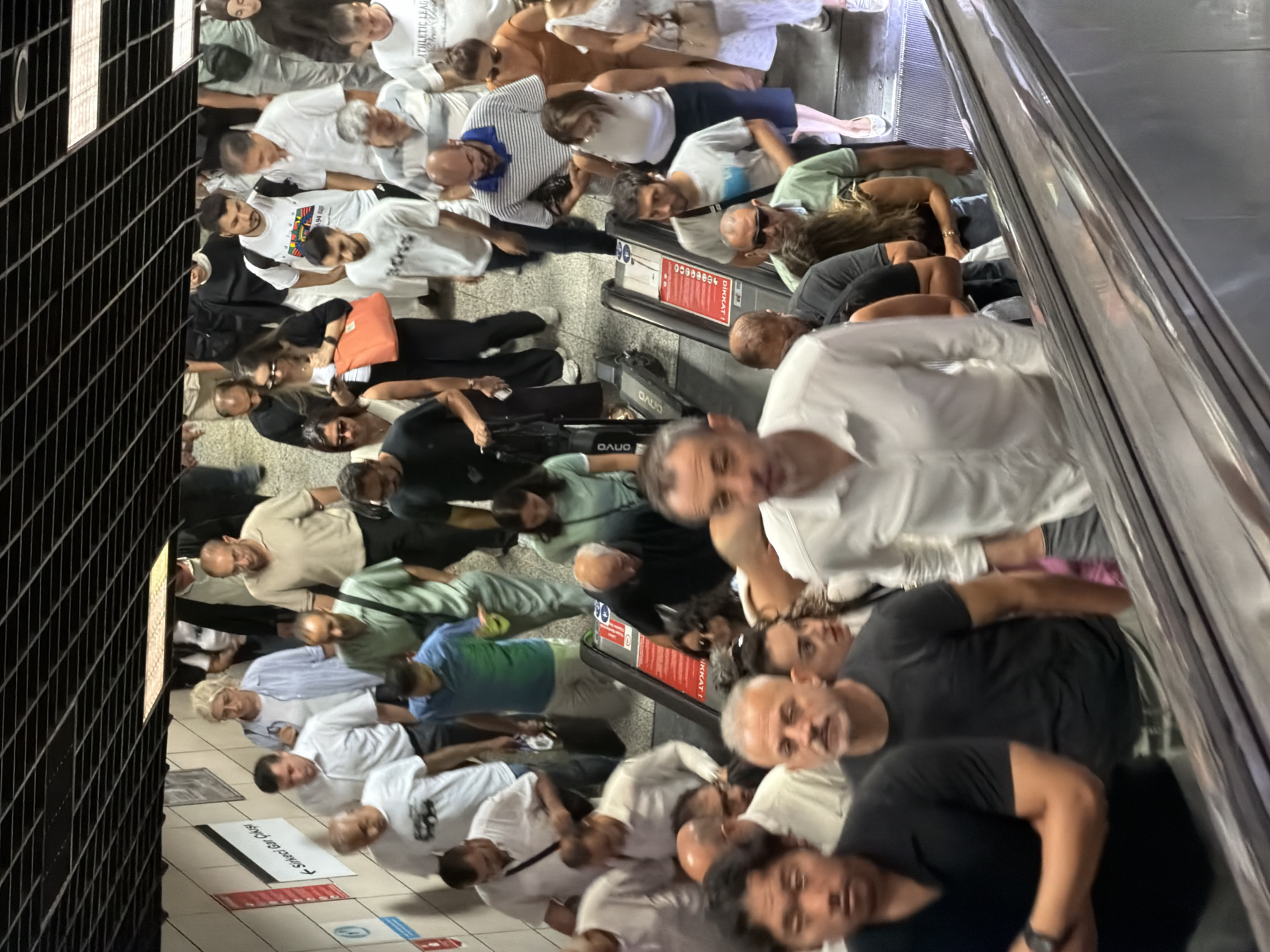 The image depicts a crowded escalator in a subway station in Istanbul, Turkey. Numerous people are ascending the escalator, most of them appearing to be commuters. The scene is captured from a high angle, looking down the escalator, with the ceiling of the station visible at the top.
The people are dressed in casual clothing, reflecting a warm climate. They are of various ages and ethnicities, and their expressions range from neutral to somewhat hurried. Some individuals are carrying bags or personal belongings. There are also two electric scooters parked on the side of the escalator. The station environment is functional and modern, with tiled walls and signage.
The overall atmosphere suggests a busy public transportation hub during a peak time. The context of the subway station, combined with the style of signage and the presence of a large number of people, strongly indicates a location within the bustling city of Istanbul.