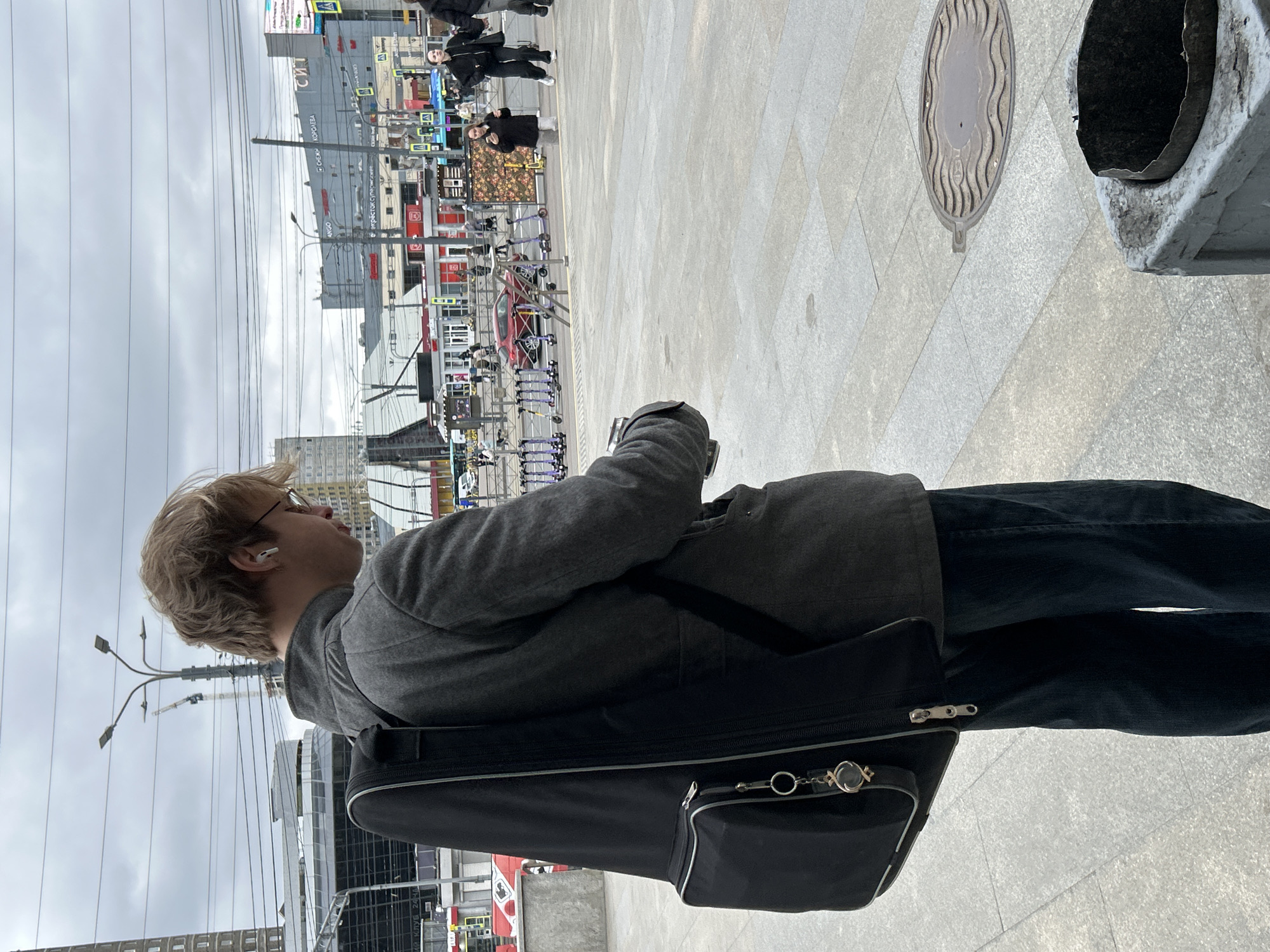 The image captures a person walking outdoors in an urban setting, viewed from their left side. The subject has blond hair, wears glasses, a grey jacket, and black pants, with a white earbud in their left ear. They are carrying a black guitar case over their shoulder, which features a small zippered pouch and decorative keychains.
The scene is a wide paved area, possibly a square or large sidewalk, with buildings in the background under an overcast sky. Overhead power lines span the frame. A multi-armed street light is visible to the left. In the distance, a construction crane can be seen.
Several other people are present in the background, though their details are indistinct. A red car is parked, and a row of rental scooters or bikes is lined up near the buildings. On the ground, a circular manhole cover with a wavy design is visible, along with part of a grey concrete structure, possibly a trash can or bollard.
Visible text on signs and buildings includes "СИГ" (SIG), "КОРОЛЕВА" (KOROLEVA), and "INGO". Partially legible text like "КЛУБ 24Ч" (CLUB 24H) also appears. The Cyrillic script and the provided context confirm the location as Saint Petersburg, Russia. The weather is overcast, indicating daytime.