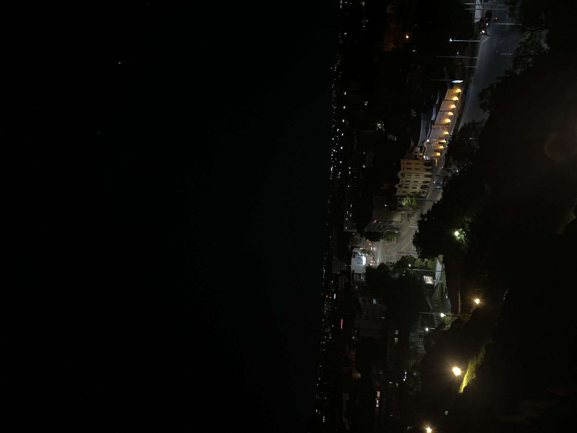 The image depicts a nighttime view of Plovdiv, Bulgaria, likely taken from a high vantage point. The left side of the image is dominated by darkness, presumably a body of water. The right side showcases the cityscape illuminated by streetlights and building lights. A distinct architectural feature, a series of arched structures illuminated with yellow lights, is visible. Streetlights line a road adjacent to the arches. Farther into the city, building lights create a speckled pattern. The overall impression is a contrast between the dark water and the illuminated urban landscape.