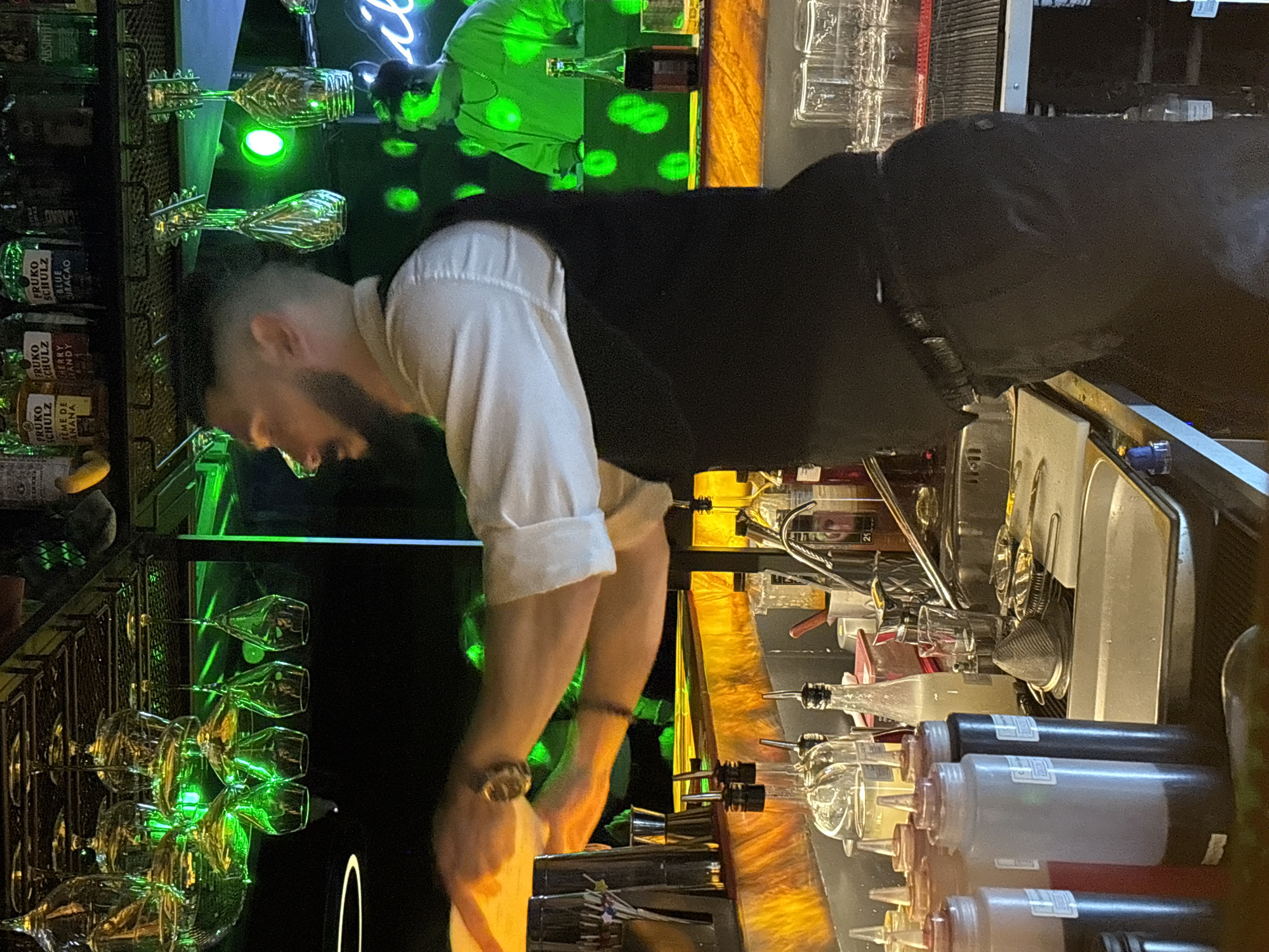 A man with a beard, wearing a white shirt and black vest, stands behind a bar. The bar is illuminated with green lighting and contains glassware, rows of bottles, and condiment dispensers. The location appears to be a bar, possibly in Perm, Russia.