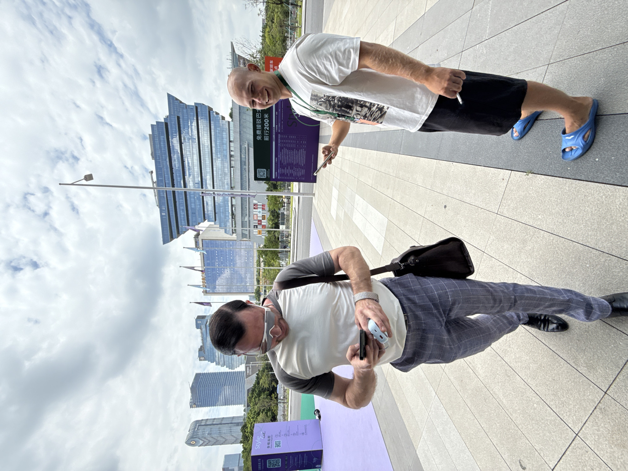 Two men are standing on a sidewalk in front of modern glass buildings, with a cloudy sky overhead. The man on the left, wearing a grey and white raglan t-shirt, plaid pants, and black dress shoes, is looking down at his phone and holding a small blue power bank. He is also carrying a dark brown messenger bag. The man on the right, wearing a white t-shirt, black shorts, and blue slides, is smiling and looking at the camera while holding his phone and a cigarette. Behind the men is a large purple sign with Chinese text and graphics. The buildings in the background are tall and modern, with reflective glass facades. The scene appears to be in an urban environment, possibly a business district or event venue in Haizhu District, China.
