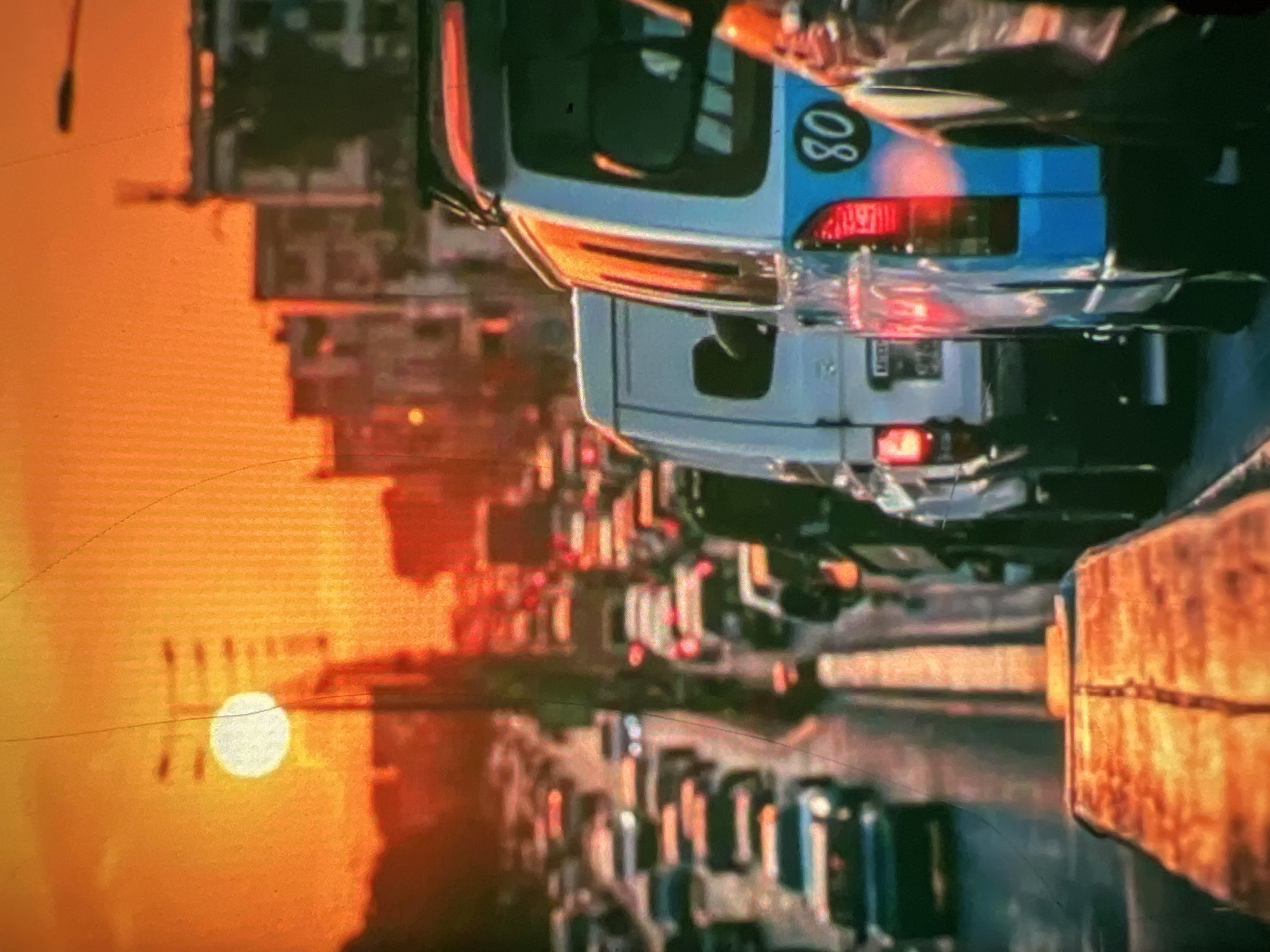 The image depicts a densely packed traffic scene in Lagos, Nigeria, during sunset. The sky is a vibrant orange, with the sun appearing as a bright, white disc partially obscured by utility poles. Buildings, appearing somewhat blurred due to the distance and lighting, line the sides of the road, suggesting an urban environment.
The road itself is congested with numerous vehicles, primarily cars and vans, moving slowly or stationary. Several vehicles are visible in the foreground, including a light blue van with the number "80" prominently displayed on its rear. This van has its brake lights illuminated, indicating it is stopped or slowing down. Adjacent to it is a white van, also appearing to be part of the traffic jam. Further down the road, more cars are lined up, their headlights and taillights creating streaks of light in the dusky atmosphere.
The overall impression is one of a typical, busy evening commute in a large African city, characterized by heavy traffic and the dramatic natural lighting of a sunset. There are no clearly discernible individuals in the image, with the focus being on the vehicles and the urban landscape. The perspective seems to be from a vehicle within the traffic, looking forward.