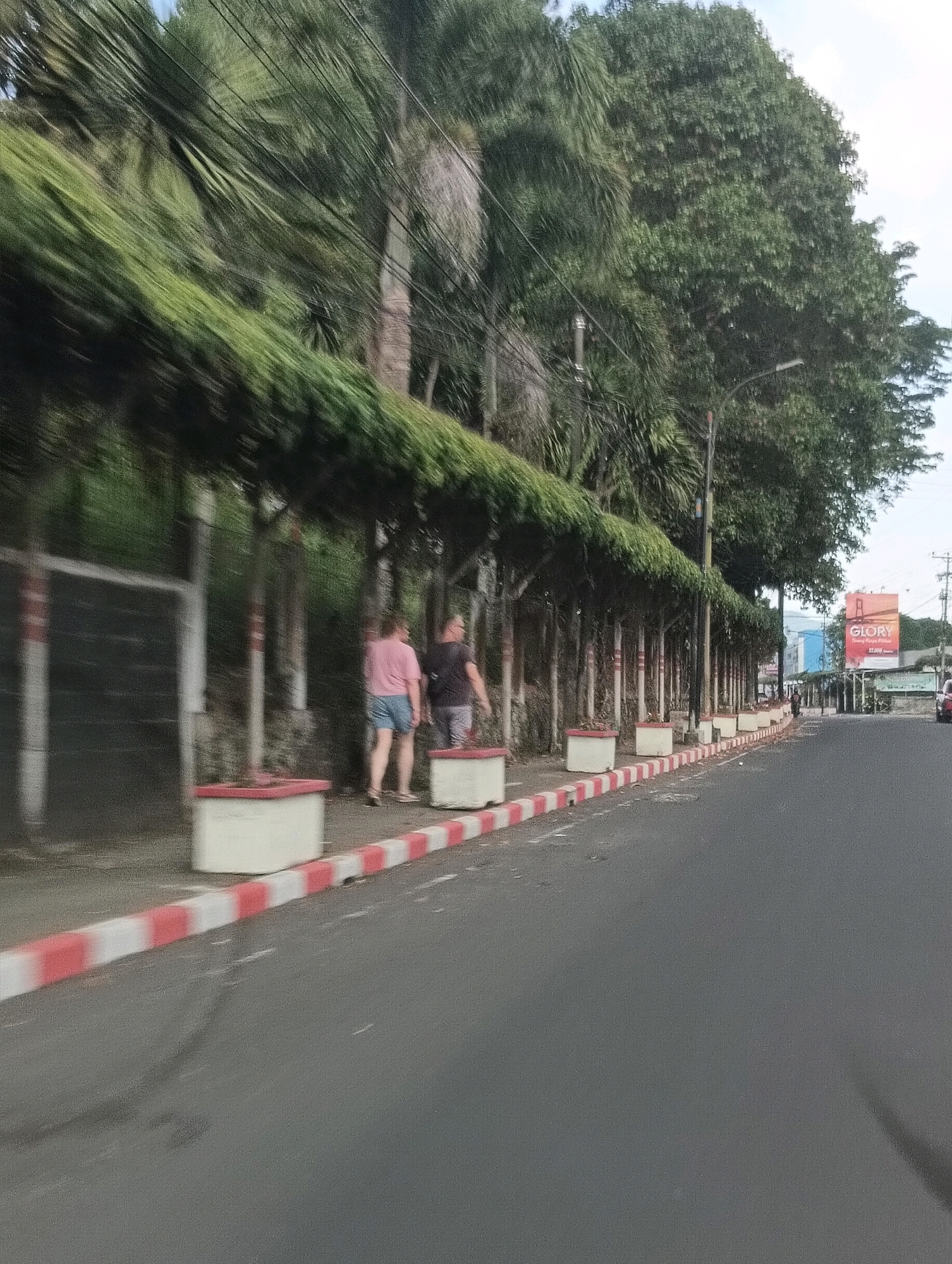 A street scene in Tomohon, Indonesia, is depicted, featuring a sidewalk lined with planters and trees. Two individuals, one wearing a pink shirt and shorts, the other a dark shirt and shorts, walk along the sidewalk. The road is paved, with a red and white striped border separating it from the sidewalk. Buildings and a billboard are visible in the background. The image has a slightly blurred quality, suggesting movement.