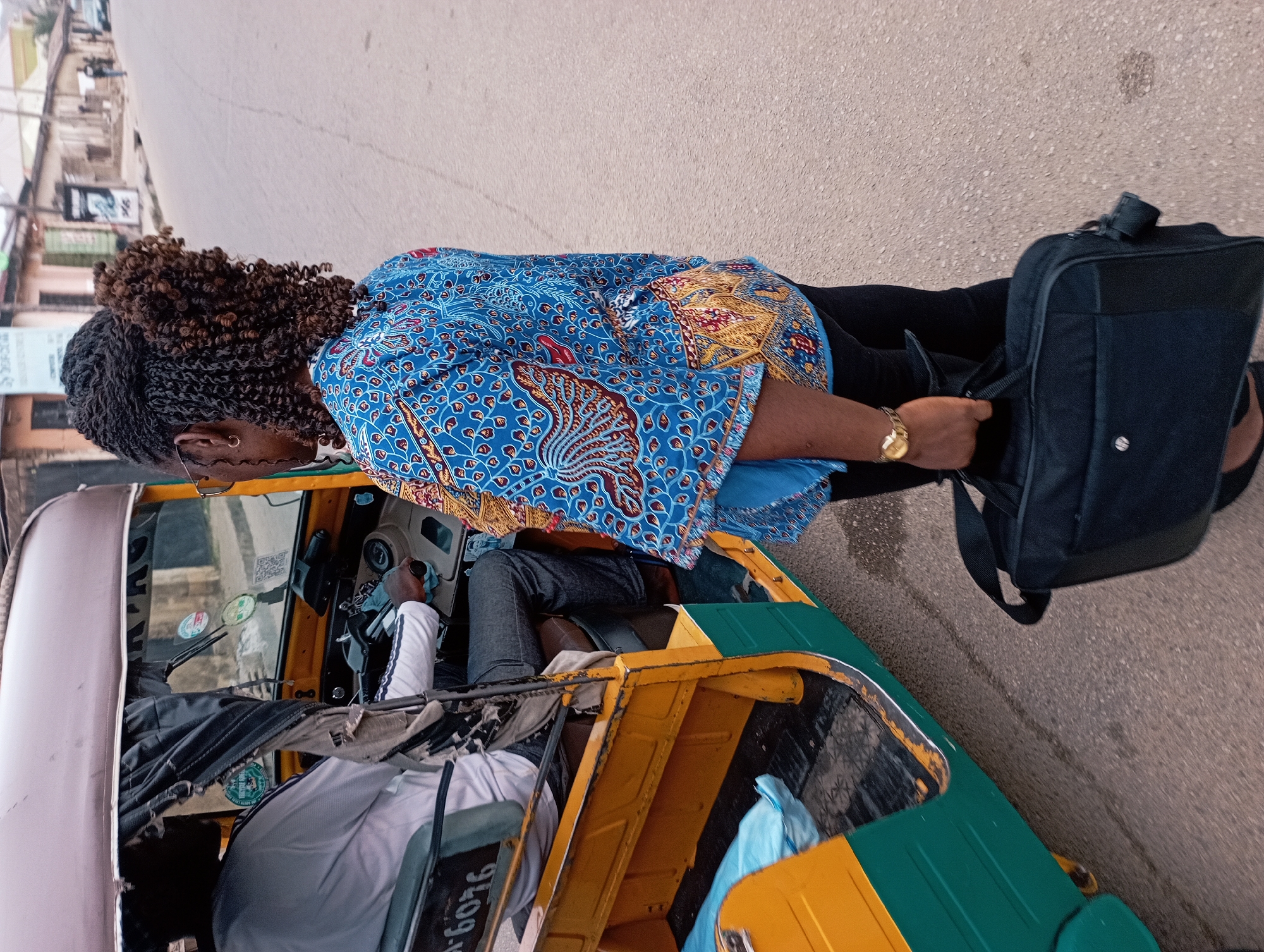 The image, oriented on its side, depicts an outdoor street scene during the daytime, likely in an urban setting. The main subject is a person, viewed from behind, standing next to a yellow and green tricycle, often referred to as a "Keke Napep" in Nigeria. This individual is wearing a distinctive blue top with intricate red, gold, and lighter blue patterns, black trousers, and has dark, curly braided hair. A gold wristwatch is visible on their left wrist, and a small hoop earring is in their left ear. They are holding a black laptop bag or backpack with their right hand.
Inside the tricycle, a driver, wearing a white long-sleeved shirt and dark trousers, is visible at the controls. The tricycle's front windshield, dashboard area, and part of its interior, including a grey seat cover with partially visible text "...6096", are shown. The vehicle itself features yellow bodywork with green sections on the lower sides and roof.
The background shows a paved road or street receding into the distance, flanked by blurry buildings and street furniture, suggesting a bustling environment. The bright natural light indicates it is daytime with clear weather. The vehicle type and setting are consistent with daily street life in a Nigerian city like Jos.