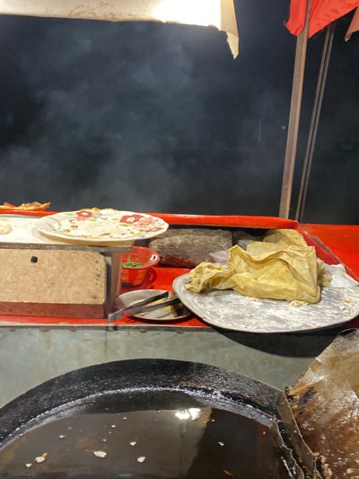 The image shows a street food stall in Kandahar, Afghanistan. The stall features a large, round pan filled with oil, likely for frying. On the stall's surface, there is a stack of flatbreads dusted with flour on a metal tray, indicating they are ready to be cooked. Additional items on the stall include a plate with partially eaten food, a chopping board, a stone weight, a small container with chopped vegetables or herbs, and a pair of metal tongs. Smoke is visible in the background, possibly from the cooking process. The stall has a red and white canopy. The overall setting suggests a busy, active street food scene at night.