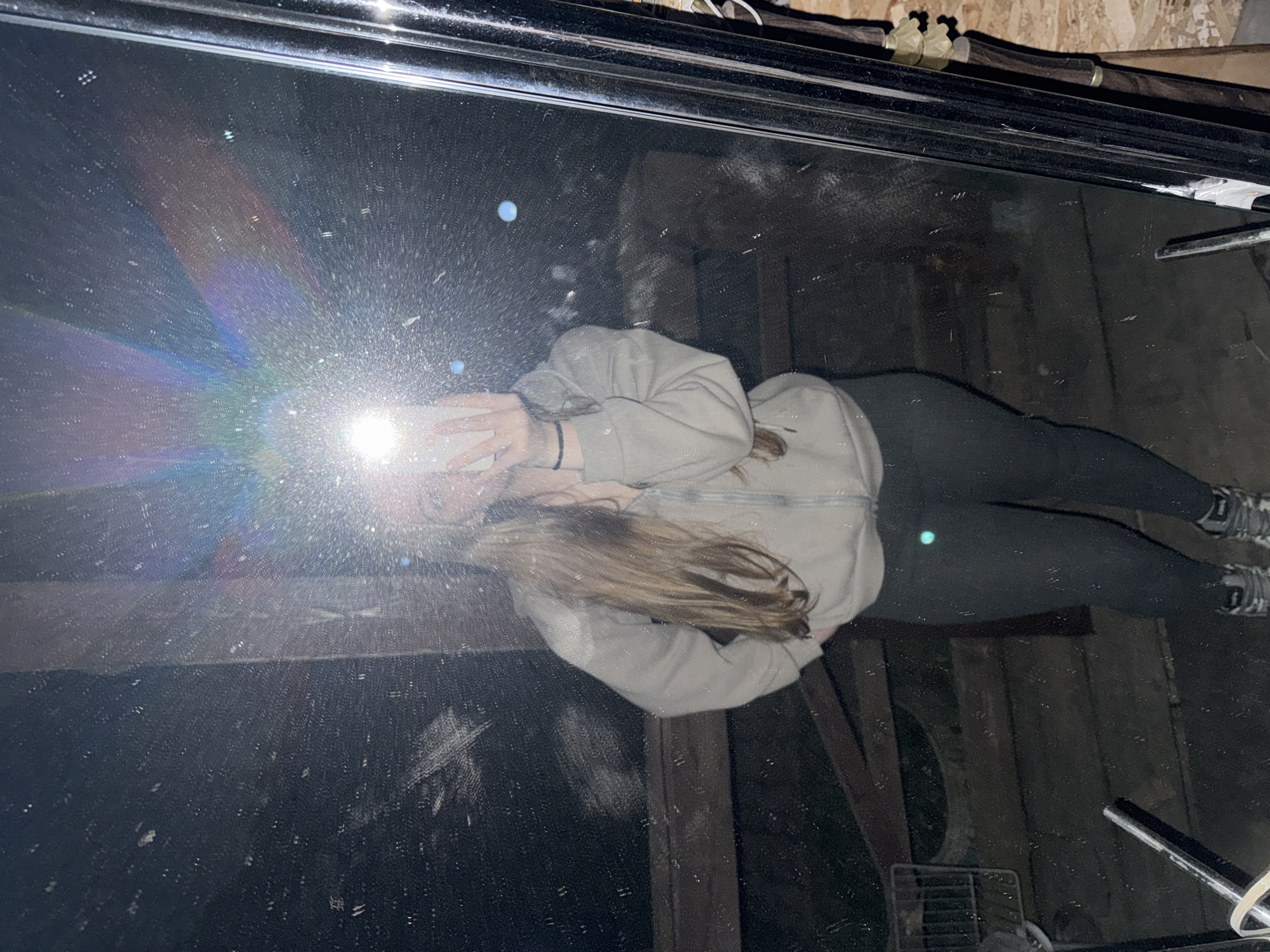 A person is taking a selfie in a mirror. The person has blonde hair and is wearing a light-colored hoodie and dark-colored leggings and sneakers. The mirror is dirty and reflects the surrounding environment, which appears to be a cluttered space with wooden walls and various objects. The flash from the phone creates a bright glare in the mirror. The exact location in Zhigalki, Russia cannot be determined from the image.