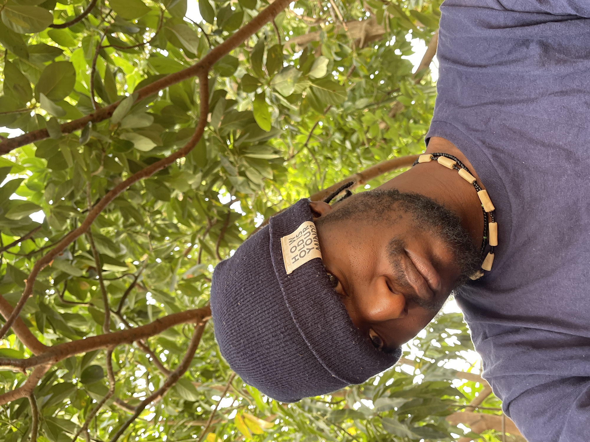 This image, rotated 90 degrees counter-clockwise, features a man captured from a low-angle perspective, looking downwards. He has dark skin, a neatly trimmed beard, and is wearing a dark blue long-sleeved shirt. A dark blue, ribbed knitted beanie covers his head, and a multi-stranded necklace with small black beads and larger, light-colored cylindrical wooden beads is visible around his neck.
The background is a vibrant canopy of green tree leaves and branches, through which bright sunlight filters, creating a luminous, dappled effect. This suggests a clear, sunny day in an outdoor, natural environment. On the folded brim of his beanie, a light-colored fabric tag is clearly visible, displaying the text "HOW OLD IS YOUR MIND" in bold lettering, followed by "SOBO" and "FOR" on subsequent lines. The man's expression is neutral and composed as he poses. The lush foliage is consistent with an outdoor setting in Jos, Nigeria.