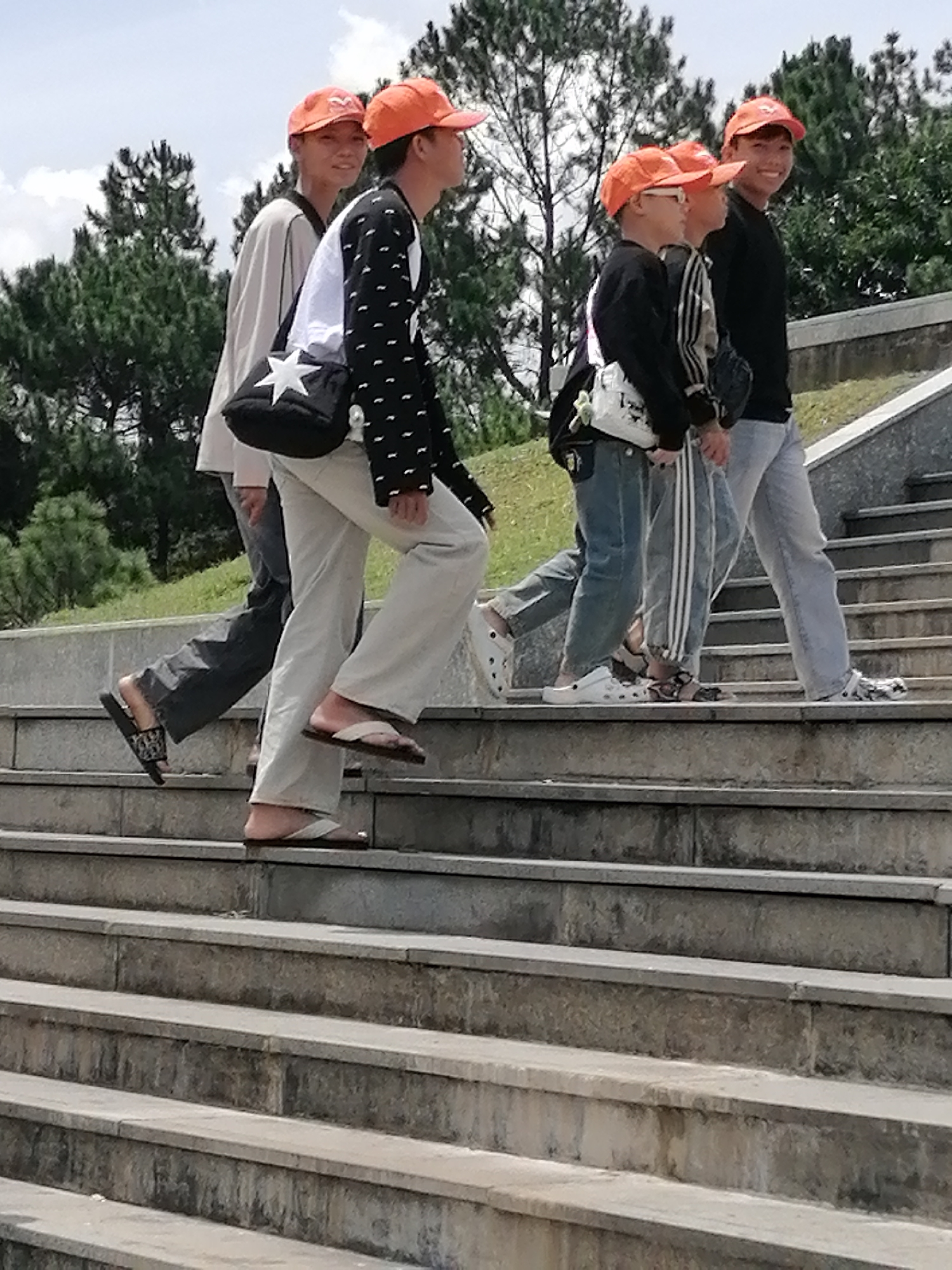 A group of five individuals, four young males and one possibly older male, are walking up a wide outdoor staircase. The staircase appears to be constructed of stone and is surrounded by lush greenery, including pine trees and grass. The sky is overcast with some visible clouds.
The individuals are dressed casually, with most wearing orange baseball caps. They are all wearing long pants and a variety of footwear, including flip-flops and what appear to be Crocs-style shoes. Several of them are carrying bags. One individual in the foreground is wearing a black long-sleeved shirt with a white pattern and light-colored pants. Another individual behind them is wearing a dark long-sleeved shirt and striped athletic pants. The individual in the far back on the right is wearing a black t-shirt and light gray pants. The person second from the left has a white bag with a black star on it.
The overall impression is that of a casual outing or exploration of an outdoor location, possibly a park or a scenic overlook, given the presence of the stairs and the natural surroundings. The location is identified as a public park in Da Lat, Vietnam, characterized by its elevated terrain and widespread use of concrete stairs in its landscaping.