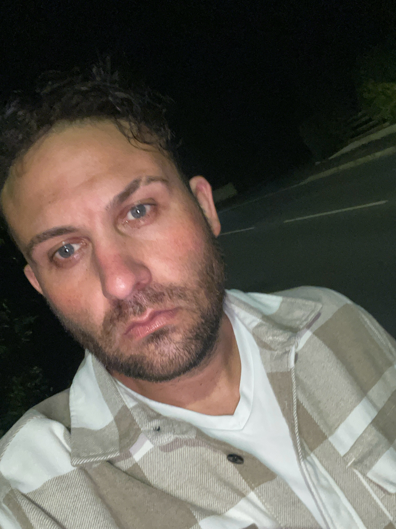 The image shows a close-up, dimly lit selfie of a person with light skin, short, dark, curly hair, and a beard. The person has light blue eyes and appears to be looking directly at the camera with a neutral to somber expression. Their face is slightly flushed. They are wearing a white t-shirt under a tan and white plaid flannel shirt. The background is dark, with a hint of greenery on the left and a paved surface with white lines on the right, possibly a road. The overall lighting suggests it is nighttime. The specific location in the city of Armagh, United Kingdom cannot be determined from the visual information provided in the image.