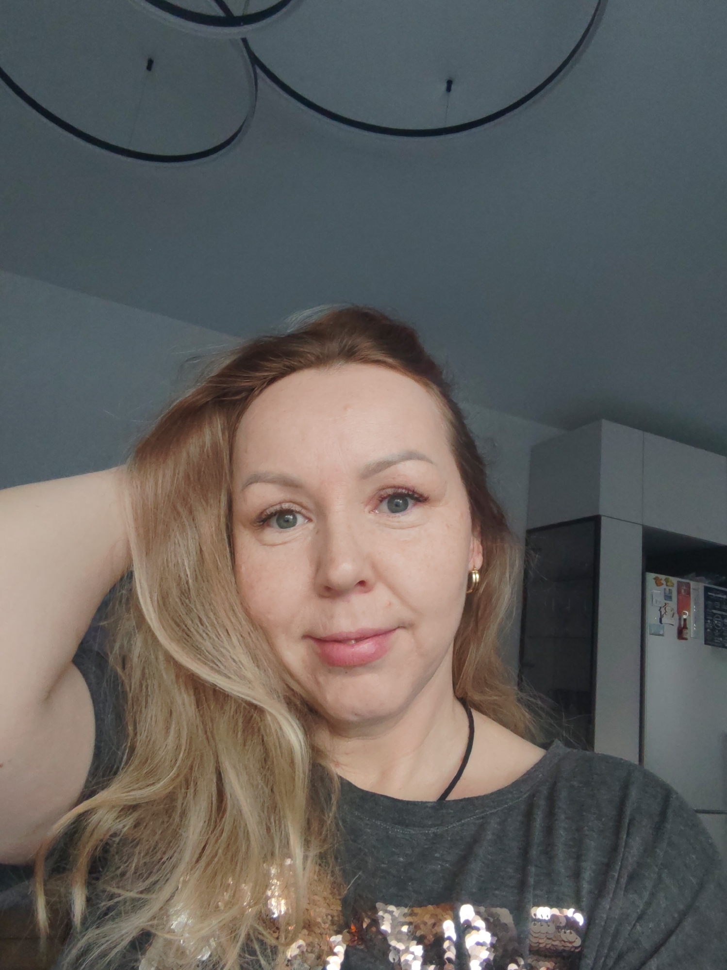 A middle-aged Caucasian woman is posing for a selfie indoors. She has fair skin, blonde hair with darker roots, and blue eyes. She is wearing a dark-colored t-shirt with sequin embellishments on the front. A thin black necklace is visible around her neck, and she has gold hoop earrings. Her left arm is raised, with her hand touching her hair.
The background includes a white wall and a modern light fixture on the ceiling. In the right side of the frame, a white cabinet or refrigerator is visible, with a few papers or magnets attached to it.
The location is the city of Chaikovsky, Russia.