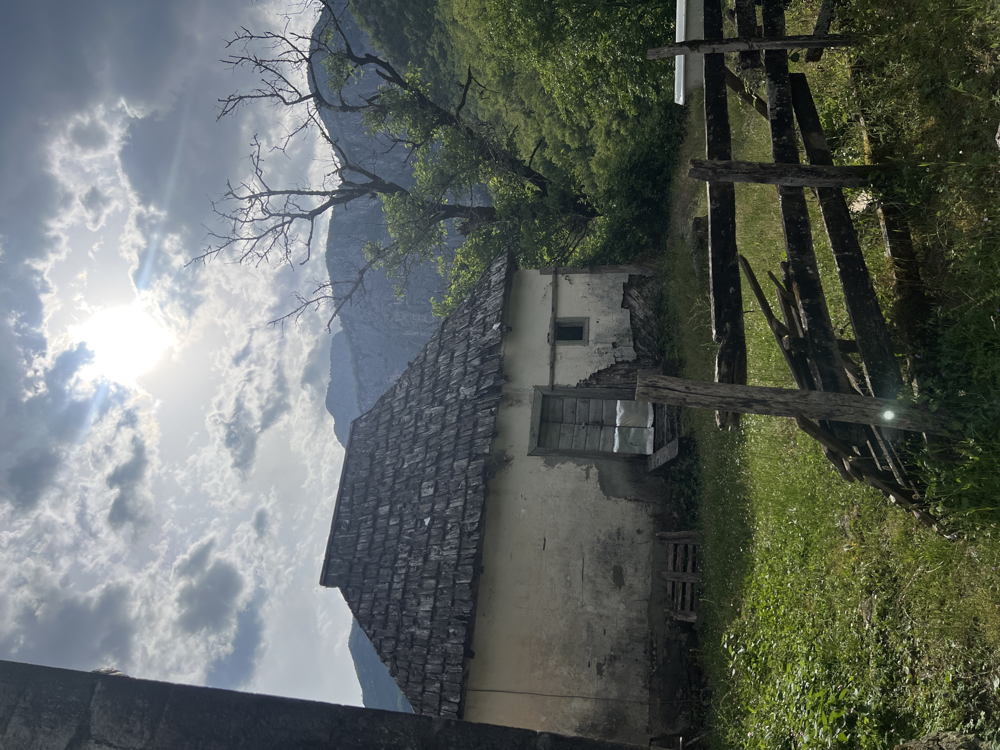 The image shows a dilapidated, one-story building with a gray tile roof in Međuriječje, Montenegro. The building has peeling white stucco walls and a boarded-up window. Adjacent to the building is a weathered wooden fence. Behind the building are green trees and a mountain range. The sky is cloudy, and the sun is visible. The foreground includes grass and parts of a stone structure.