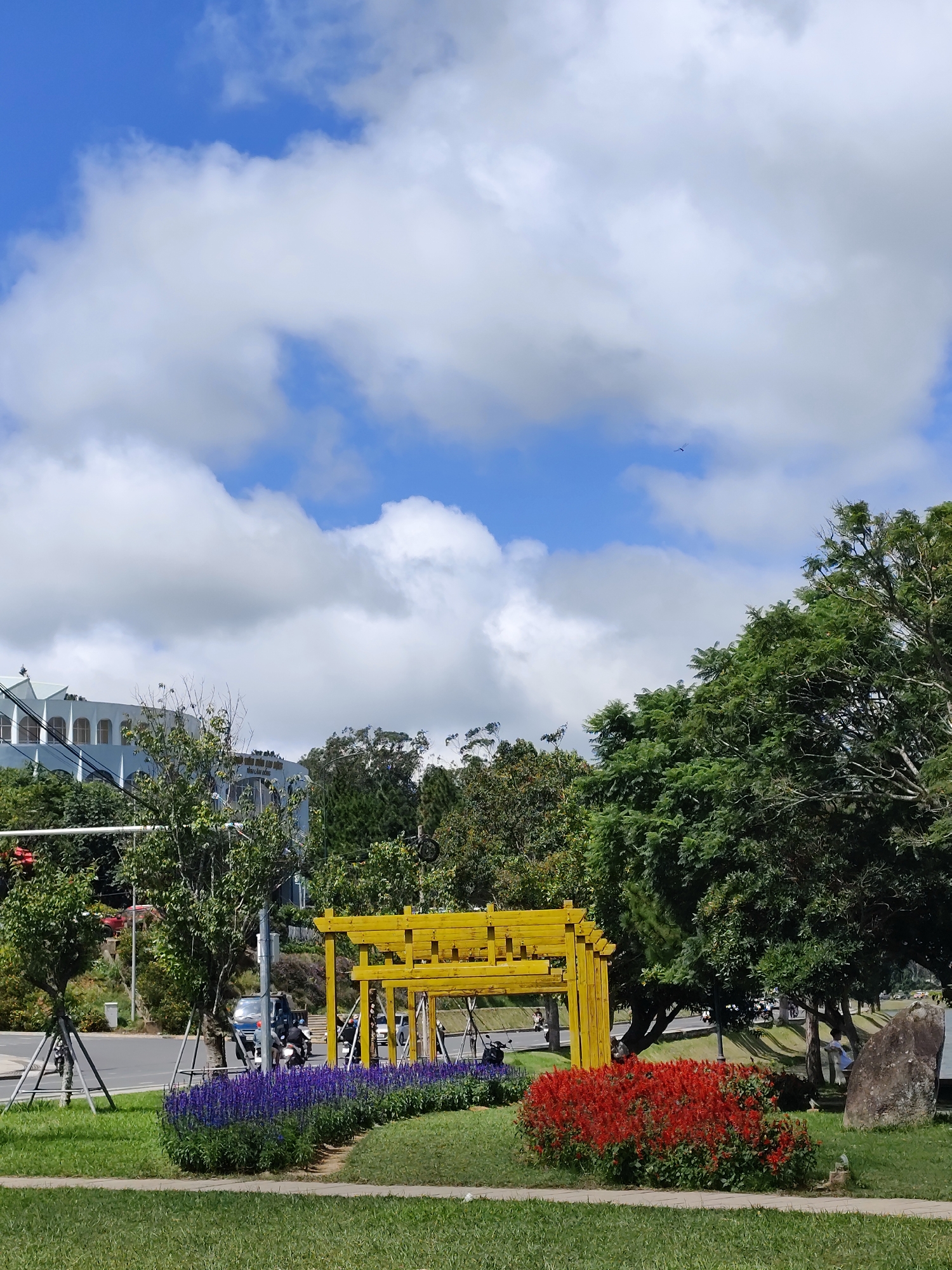 This image depicts a bright, sunny day in Da Lat, Vietnam. In the foreground, a meticulously manicured lawn is bordered by a path and then by vibrant flower beds. A bed of deep purple flowers runs along the left side, followed by a dense patch of bright red flowers to the right. Behind the flowers, a modern yellow wooden archway structure stands, providing a central focal point.
Beyond the archway, a road is visible with several motorcycles and a car in transit, suggesting a moderately busy urban setting. To the left of the road, a large, light blue building with distinctive architectural features, possibly a hotel or public building, is partially visible behind lush green trees. The right side of the scene is dominated by mature trees with full, green canopies, casting shadows on the ground below. A large, grey boulder is positioned on the right side of the frame, adding a natural element to the landscape.
The sky is a brilliant blue, punctuated by fluffy white cumulus clouds, some of which are quite large and dramatic, indicating fair weather. A tiny bird can be seen soaring high in the sky, barely visible against the vast expanse. The overall impression is one of a well-maintained urban park or roadside attraction within the picturesque city of Da Lat.