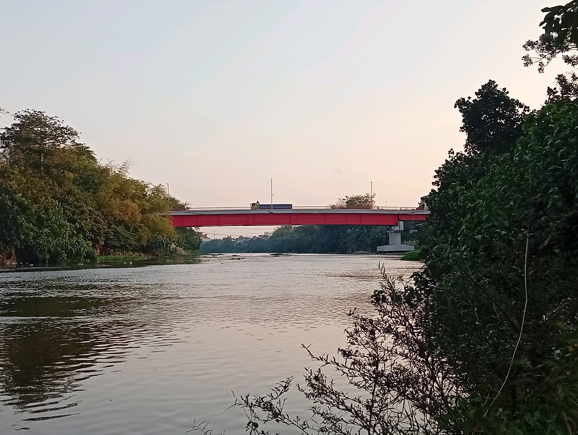 A wide, calm river flows towards the viewer, reflecting the pale sky. On the far bank, a long, red bridge spans the water, with a truck visible crossing it. Lush green trees and vegetation line both sides of the river, with some buildings visible in the distance behind the bridge. The sky is a soft gradient of light blue and pink, suggesting either dawn or dusk. This scene is likely located in Kediri City, Indonesia, possibly along the Brantas River, known for its bridges and surrounding greenery.