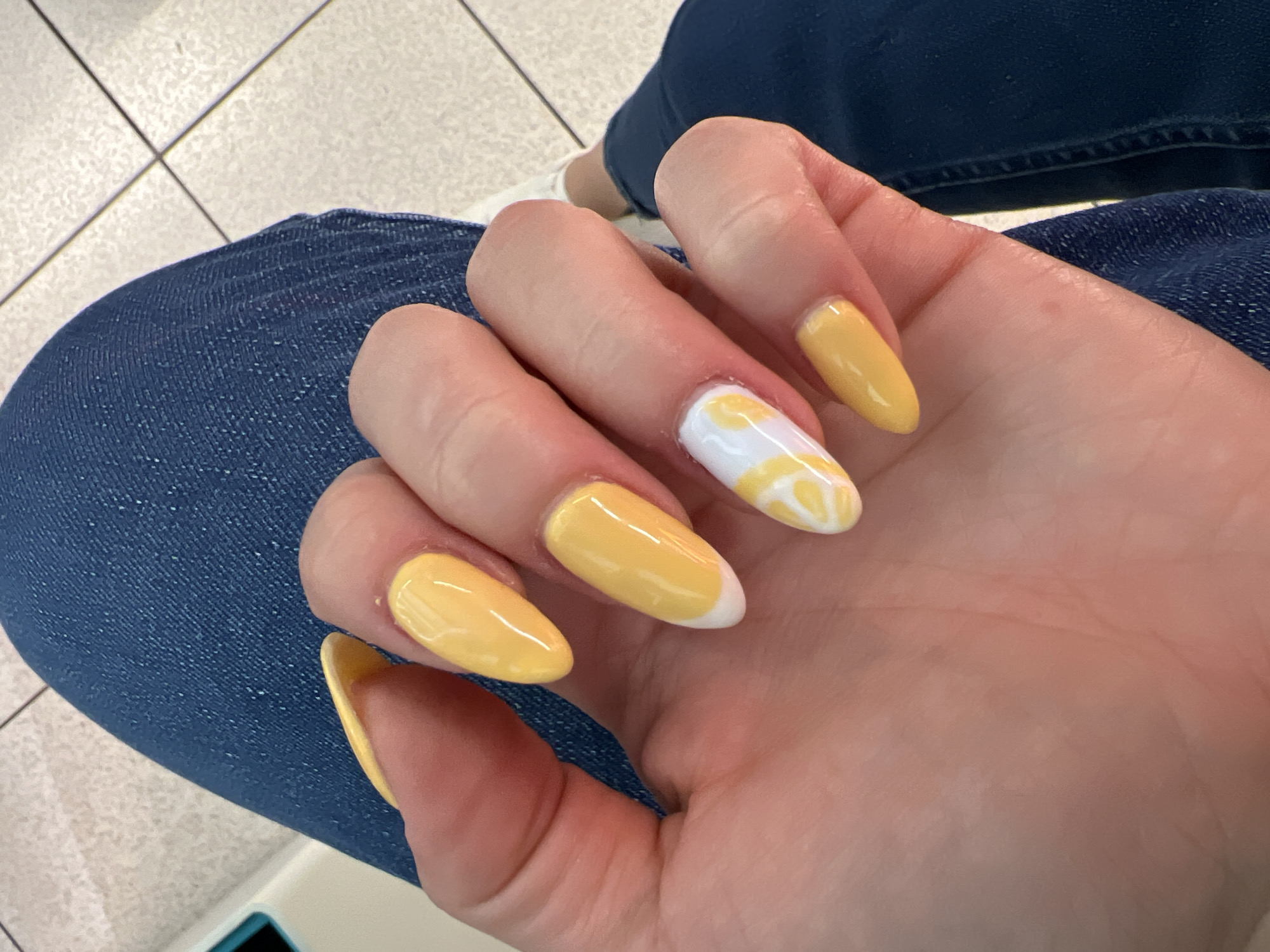 The image shows a close-up of a person's hand with bright yellow almond-shaped nails, featuring a white and yellow swirl design on one nail. The hand is resting on a person's blue denim jeans. The background appears to be an indoor tiled floor. The image does not provide any information about the city of Pazardzhik, Bulgaria.