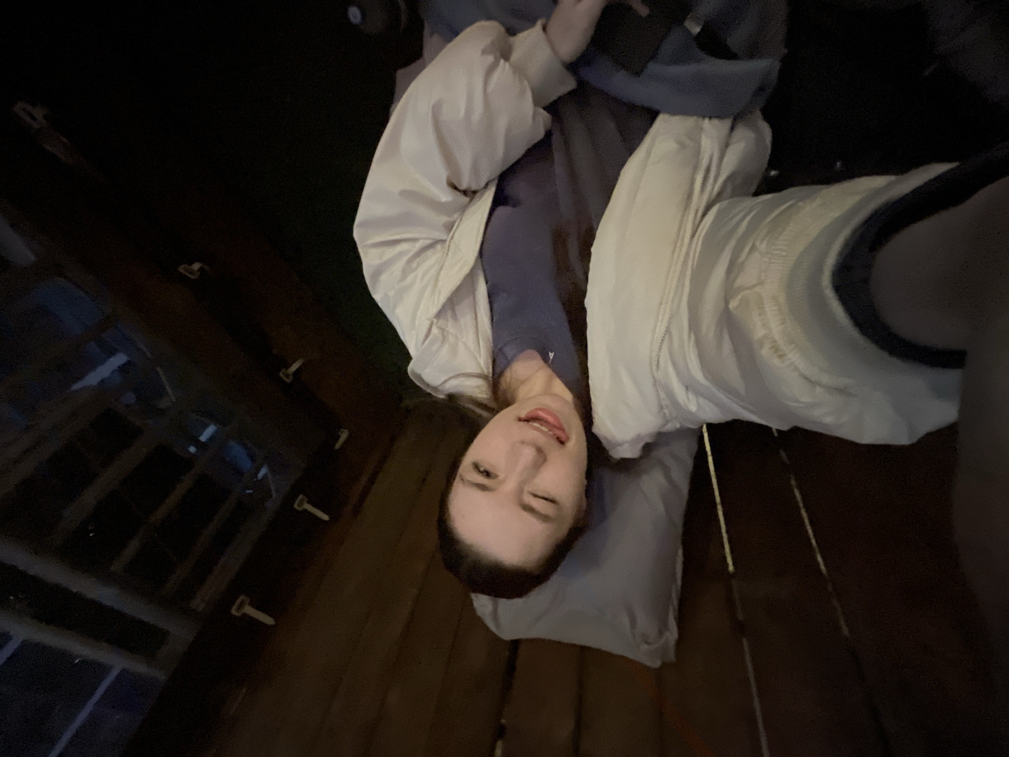 A young woman is lying on a bed in what appears to be a rustic, wooden structure, possibly a cabin or a treehouse. The structure's interior is dimly lit, with a wooden floor visible beneath the bed and wooden walls featuring rectangular window frames. The woman is wearing a light-colored jacket over a dark top. She is winking at the camera. The image is taken from an unusual angle, as if someone is standing above her looking down. It is unlikely the location can be determined to be Moscow, Russia based on the current image.