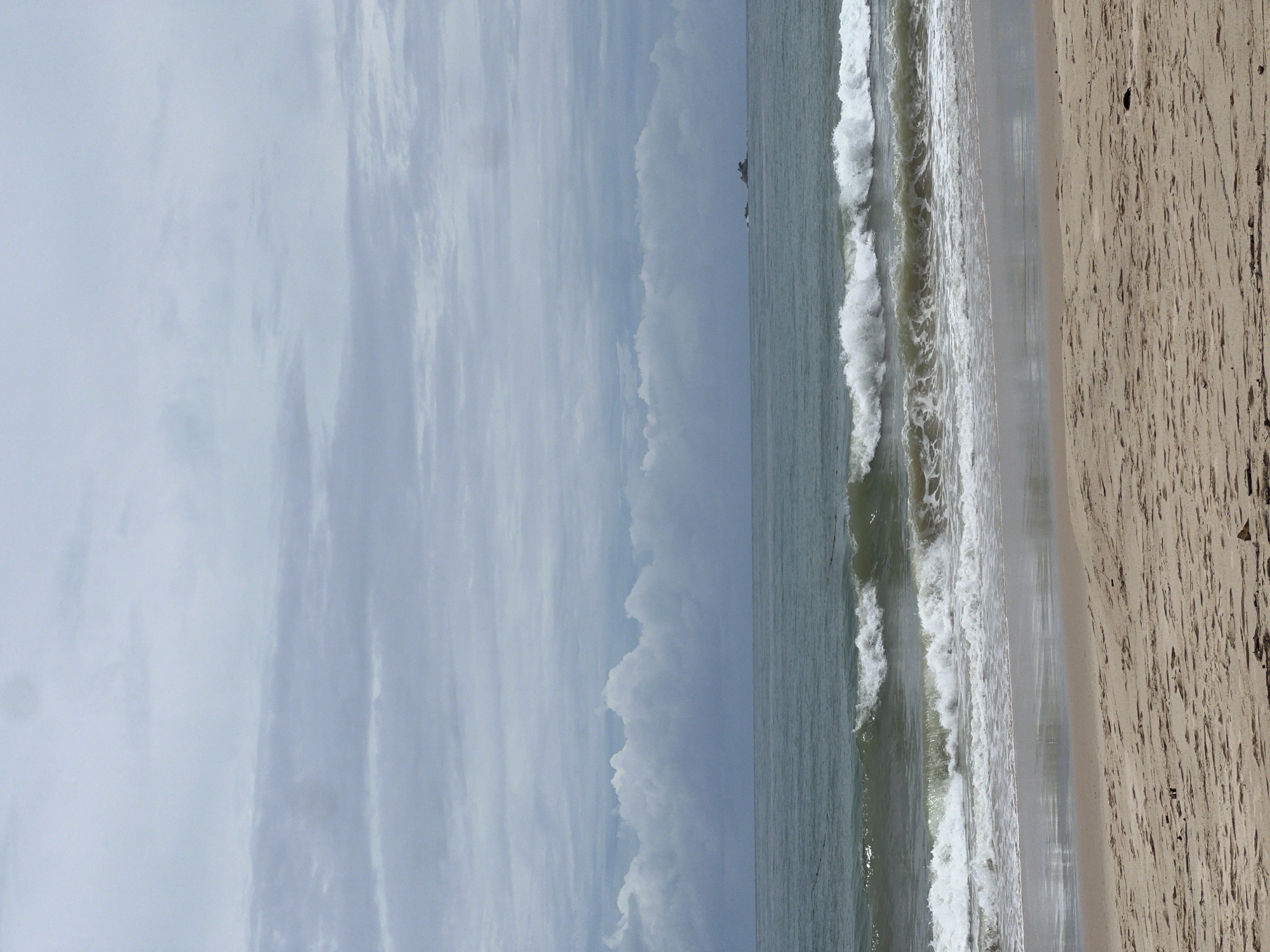 This image depicts a beach landscape in an unspecified location. The scene is divided into three main sections: the sky, the sea, and the sandy beach. The sky occupies the left side of the image, displaying a light blue color with varying cloud formations. Adjacent to the sky is the sea, where gentle waves are visible as they approach the shore. The waves create white foam as they break on the beach. On the right side, the beach stretches out with textured sand, showing patterns possibly caused by footprints or natural elements like wind and water. The overall impression is a tranquil coastal scene under an overcast sky.