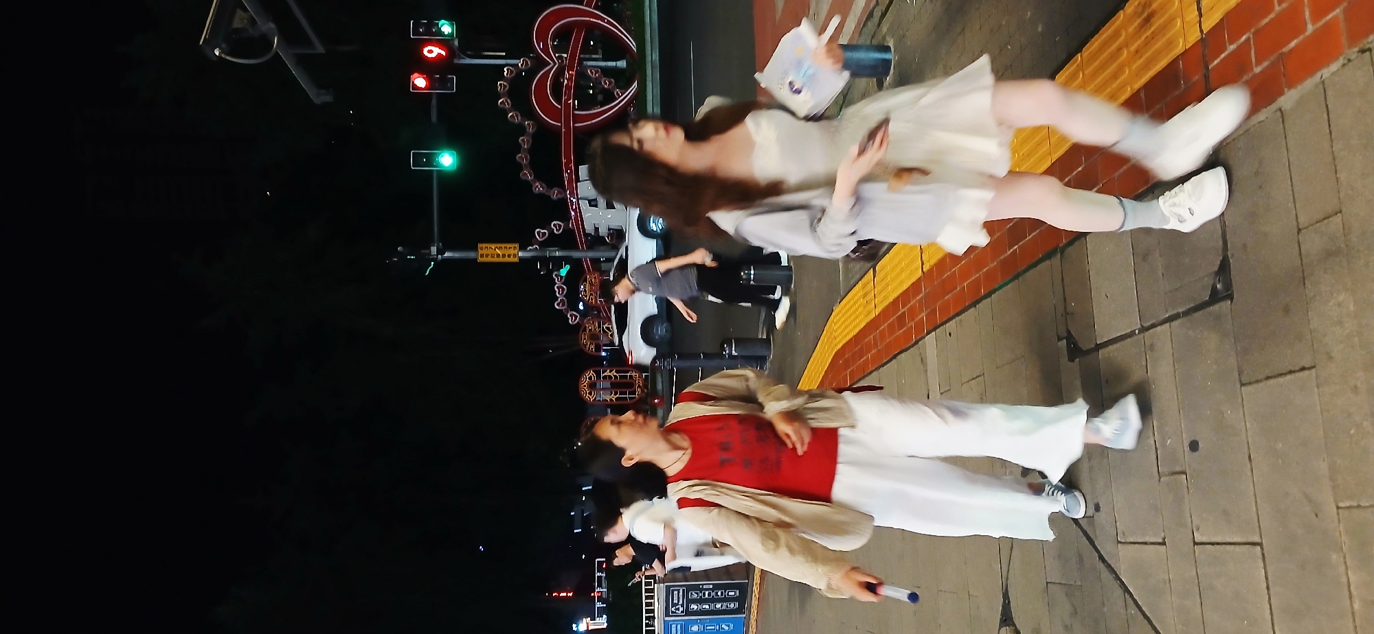Here is a street scene at night in Yuzhong District, China. There are multiple people in the image, including two women standing side by side on a sidewalk with a reddish-orange stripe. The first woman is wearing a red t-shirt, white pants, and a light brown jacket. She is holding a cylindrical object. The second woman wears a white dress and is holding a book. In the background, there is a street at night, including lit-up hearts and traffic lights.