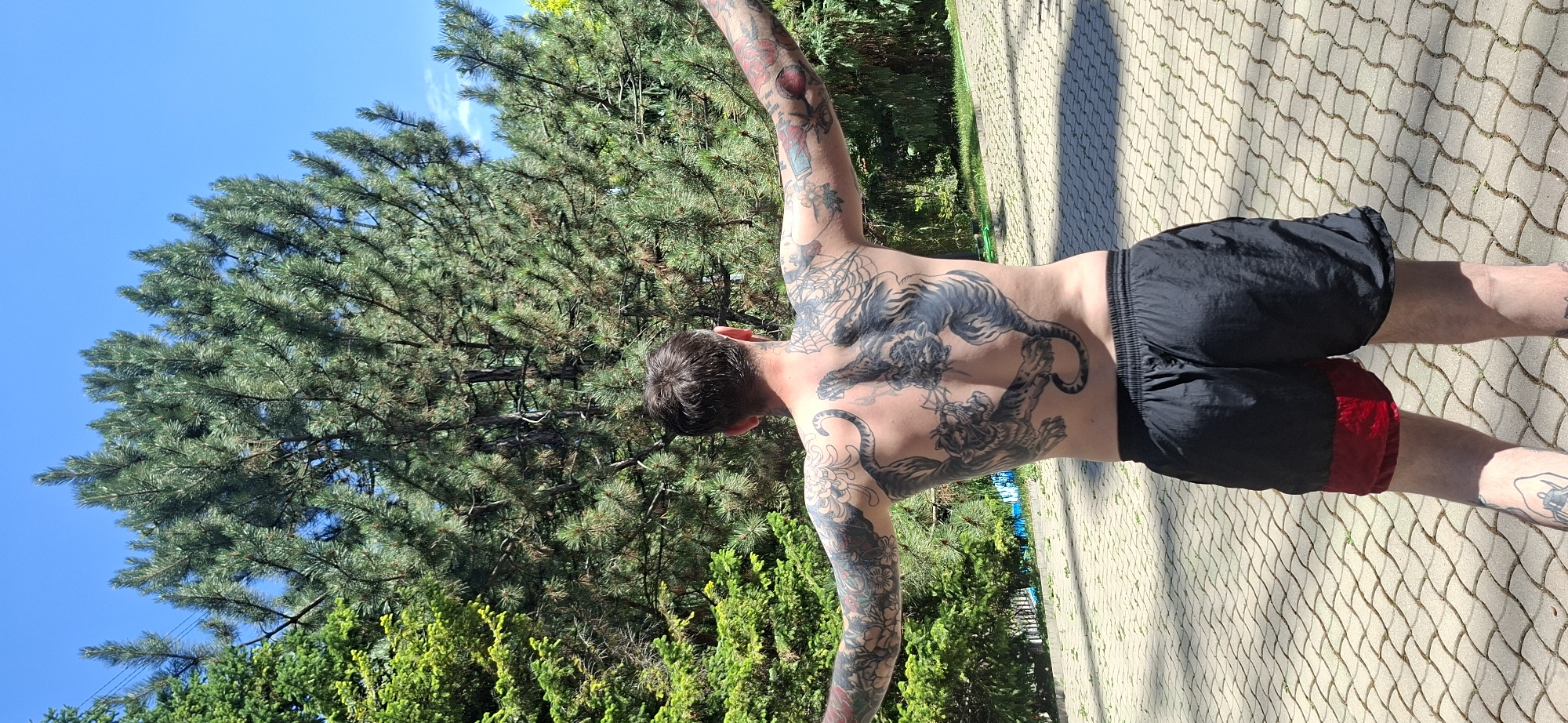 A man with extensive tattoos on his back and arms is standing outdoors with his arms outstretched. He is wearing black shorts with a red lining. Behind him, large evergreen trees and a bright blue sky are visible. The ground is paved with interlocking bricks. There is no information available in this image to suggest the location is Almaty, Kazakhstan.