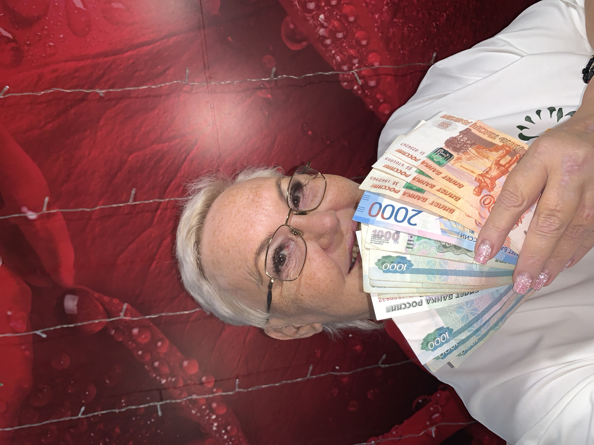 A medium shot shows a white woman with short blonde hair, wearing glasses and a white shirt, holding a fan of Russian Ruble banknotes in front of her face. The background is a red fabric with a pattern of water droplets and what appears to be decorative string lights. The woman's fingernails are painted with glitter. The image does not contain any information about the city of Chaikovsky, Russia.