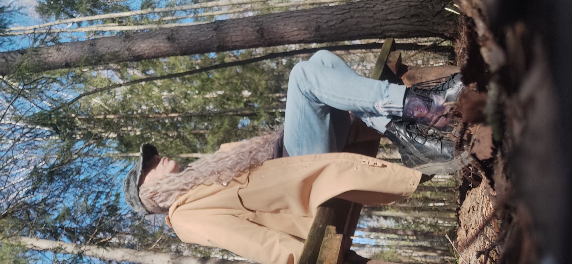 A person wearing a tan coat, blue jeans, and black boots is sitting on a wooden structure in a forest. The person is wearing a fur hat and scarf. The background consists of trees and a blue sky. The location is in a forest and its geolocation is unknown.