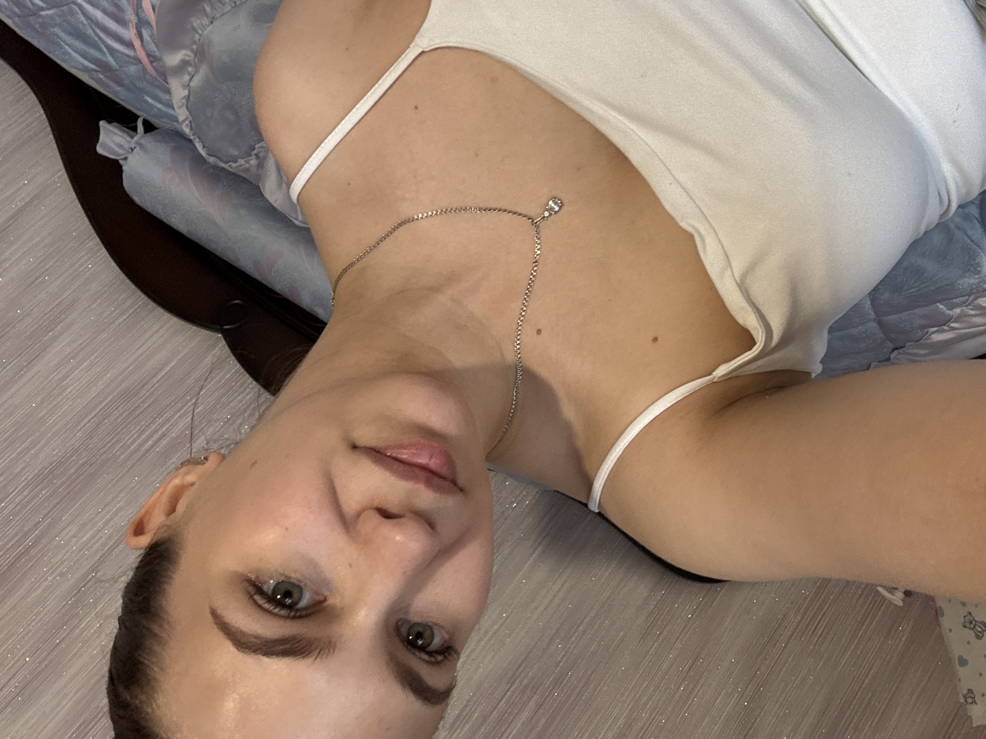 A woman with green eyes and light skin is depicted in a selfie. She wears a white tank top and a silver necklace with a small pendant. She is lying on a bed with blue patterned bedding and a dark brown headboard. The background features a light-colored wooden floor. There is no information about the location being in the city of Nizhnyaya Gar', Russia.