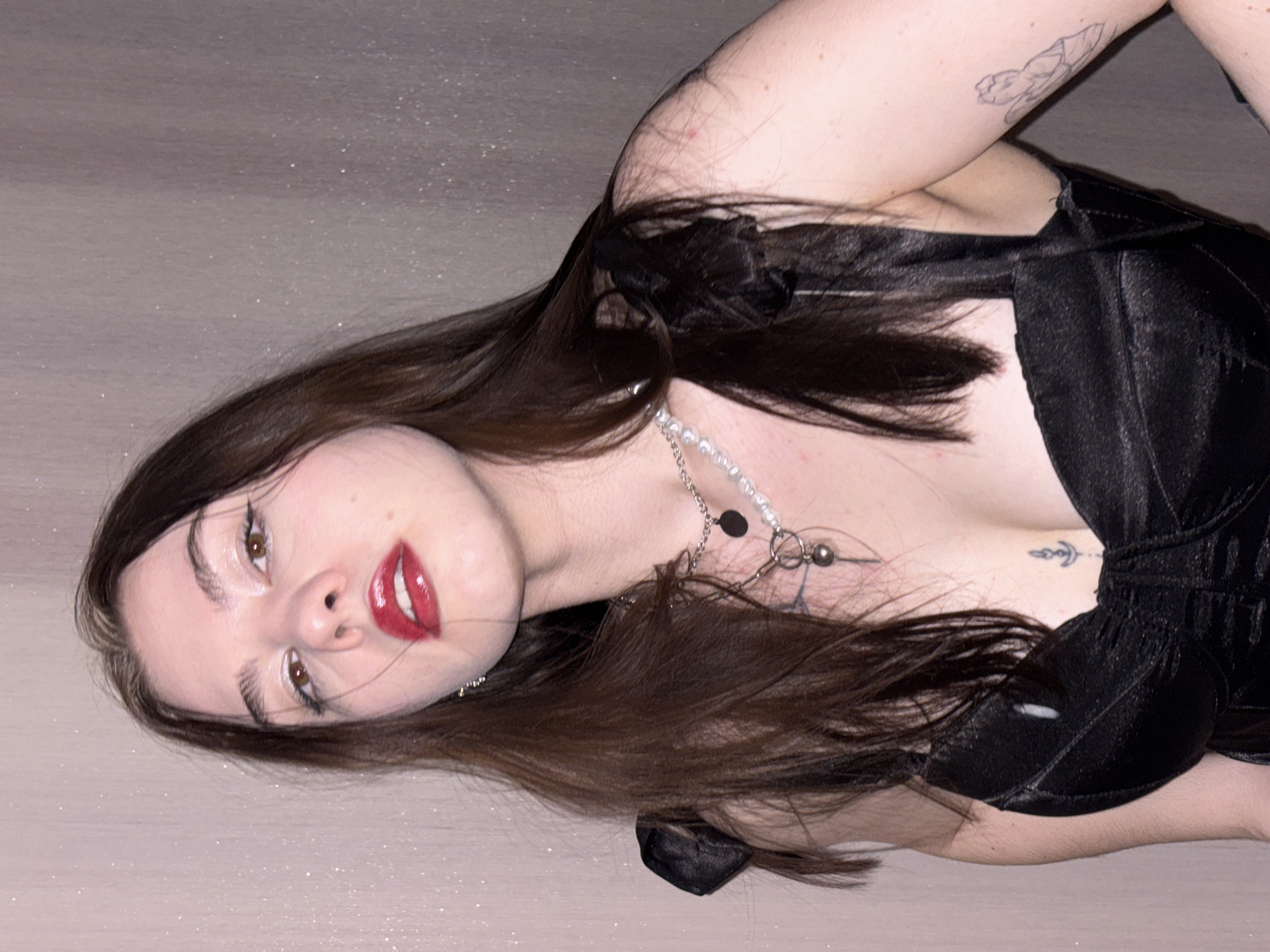 A young woman lies on a light-colored surface. She has brown hair, light skin, and is wearing a black top and a silver necklace. She has red lipstick and visible tattoos on her arm and chest. The surface she is lying on appears to have a slight shimmer. The location is unknown and cannot be determined from the image.