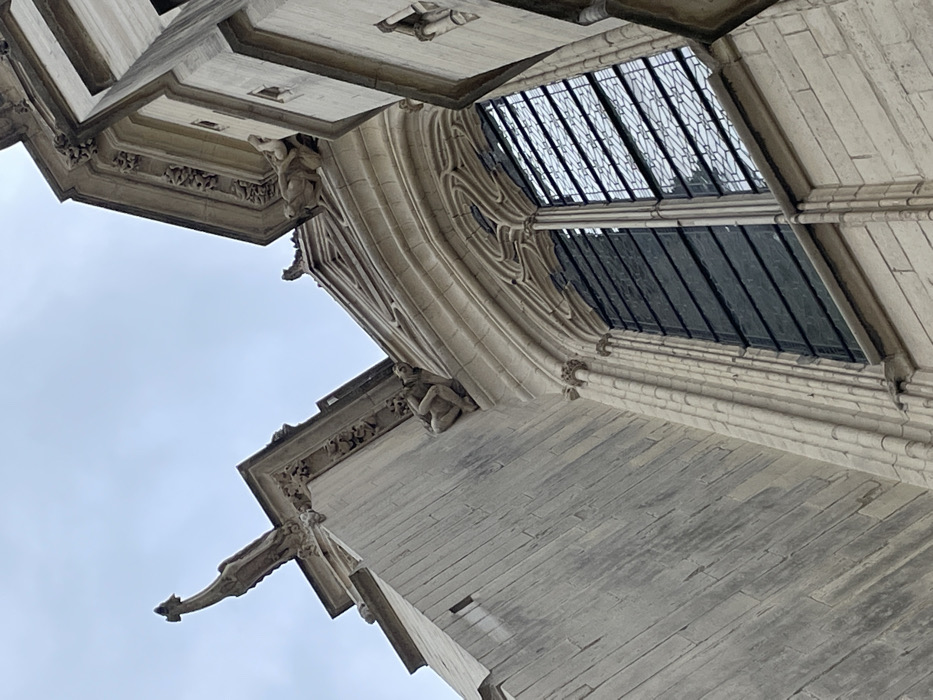 This image captures a detailed, low-angle view of the upper architectural elements of a historical stone building, identified as a section of the Sainte-Chapelle de Vincennes within the Château de Vincennes, France. The structure is composed of light-colored, aged masonry blocks.
Dominating the mid-right is a tall, arched Gothic window featuring intricate stone tracery and vertical mullions. The window's panes exhibit a mix of reflective surfaces, showing the overcast sky, and darker, possibly leaded or opaque glass sections.
Three distinct stone figures are visible on the building's exterior. On the far left, a prominent zoomorphic gargoyle with an elongated form projects outward from a corner near the roofline. Below a decorated cornice on the left facade, a crouching, humanoid figure is sculpted onto a ledge. Further to the right, above the main window and beneath another cornice, a grotesque, possibly animalistic, figure lies sprawled on a ledge.
The building features multiple ornate cornices adorned with decorative carvings, including rosette patterns, and displays architectural elements such as vertical pilasters and buttressing. The background consists of a uniform, overcast grey sky. No human subjects or active interactions are present; the scene focuses solely on the static architectural details and sculptures.