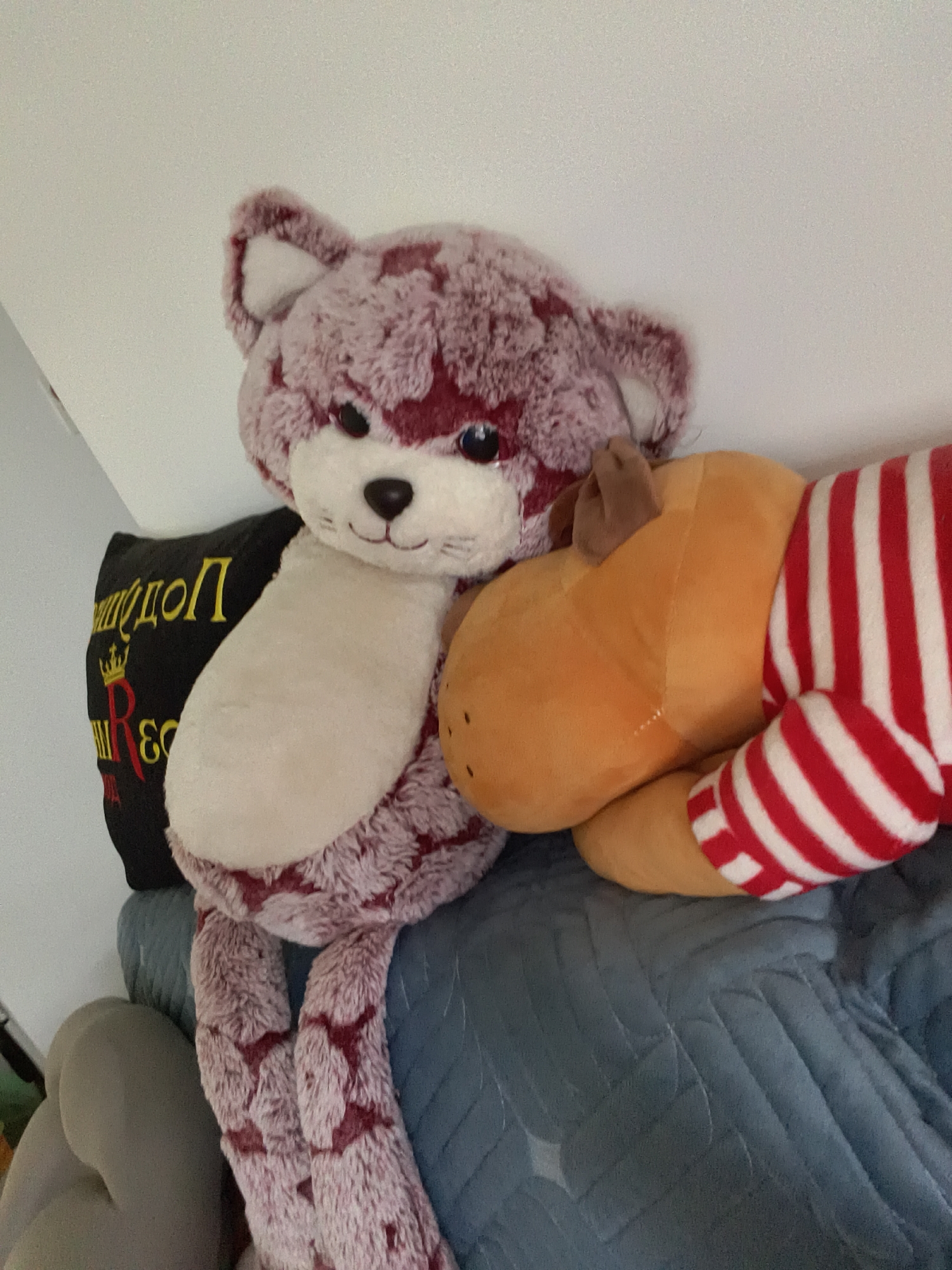 The image shows a stuffed cat and a stuffed reindeer resting on a blue quilted surface. The cat is primarily mauve and white, sitting upright with its paws dangling. To the left, there is a black pillow with gold and red lettering that seems to say "подоп" and a crown. The reindeer, wearing a red and white striped top, is nestled against the cat. The background suggests an interior space, possibly a home. There is no information to suggest the location in Petah Tikva, Israel.