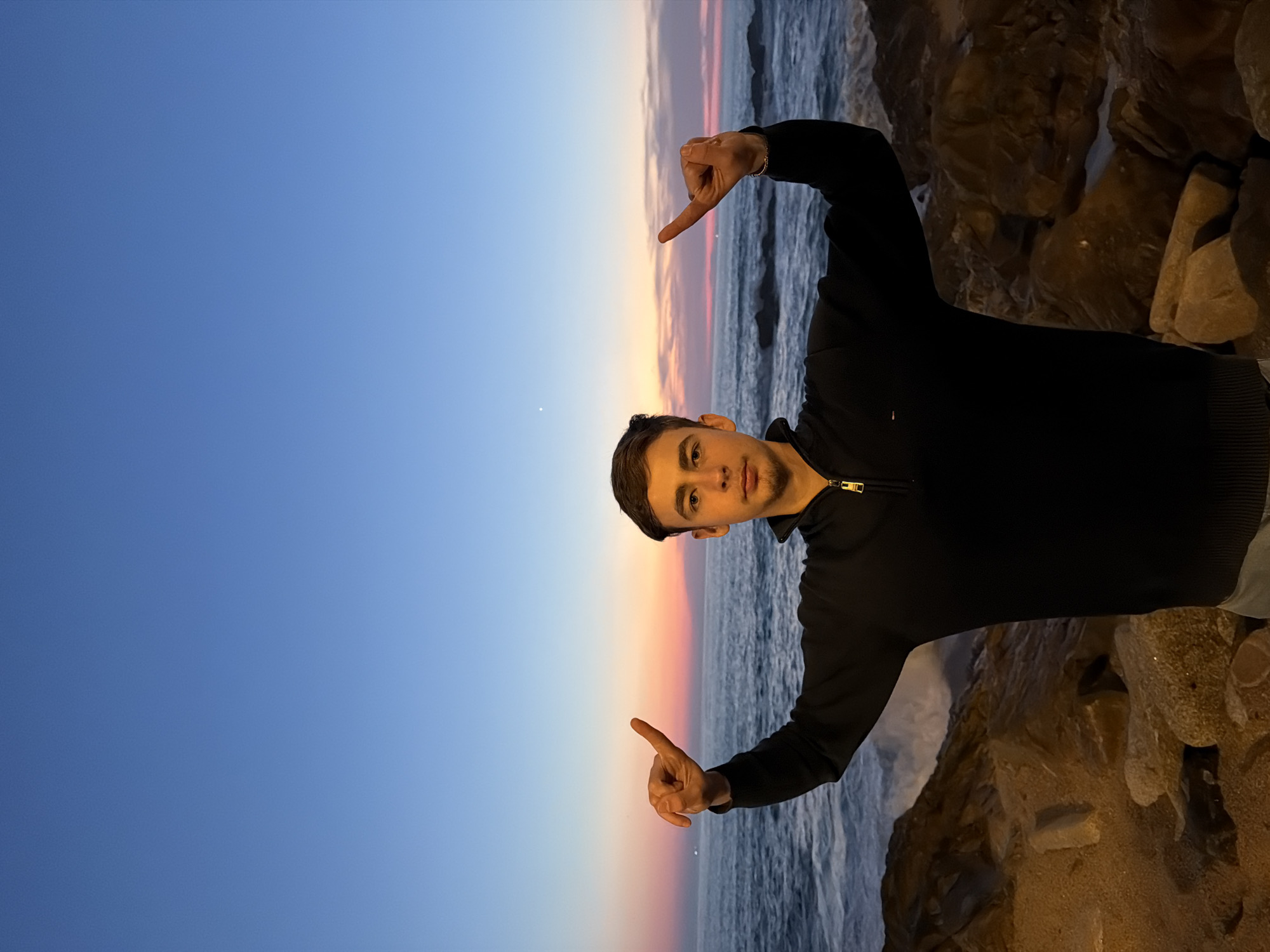 A young adult male stands centered, facing the camera, against a twilight backdrop over the Atlantic Ocean in Porto, Portugal. He has short, dark hair and light facial stubble, wearing a dark blue or black quarter-zip long-sleeved sweater with a gold zipper and a small rectangular logo on the left chest. His arms are raised, with index fingers extended outwards and slightly upwards, creating a pointing or framing gesture. His expression is neutral.
Behind the subject, the ocean surface is active with waves breaking against a rocky shoreline, visible in the lower foreground and mid-ground. The sky above transitions from a deep blue at the top to vibrant orange, pink, and light blue hues at the horizon, indicative of sunset. A single bright celestial object is visible in the upper-left sky. Faint lights are discernible on the distant horizon to the left. The scene is illuminated by ambient twilight and possibly artificial light sources casting warmth on the foreground rocks.