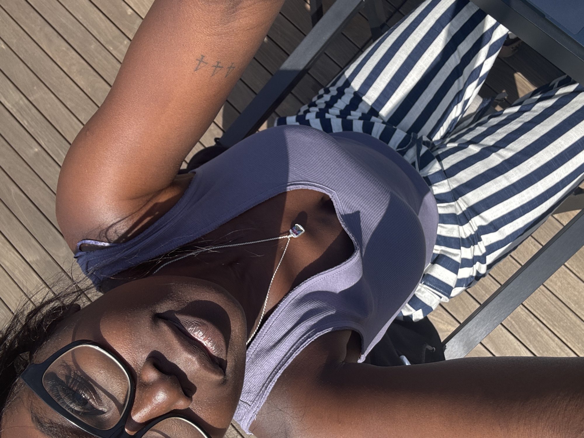 A close-up, top-down perspective captures a person seated on a chair on a wooden surface, possibly a deck or patio. The individual has dark skin, wears glasses, and a light purple tank top. A silver necklace with a pendant rests on their chest. The person is also wearing blue and white striped pants. A tattoo of small cross-like symbols is visible on their arm. The wooden surface and the chair's metal frame are also visible. The location is indeterminate but is potentially within the city of Los Angeles, United States of America.
