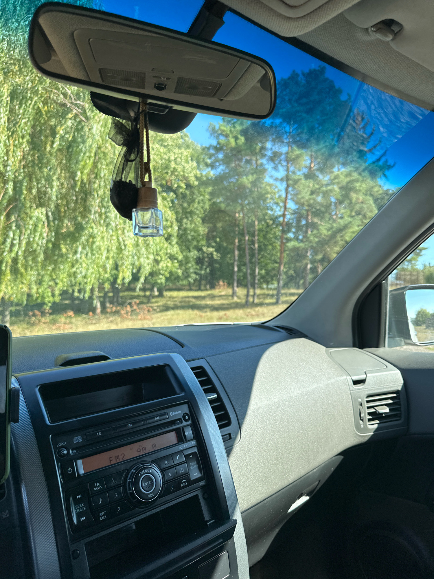 The image, taken from inside a vehicle, shows a view through the windshield of a sunlit, tree-lined area. The dashboard of the car is visible in the foreground, with a radio unit displaying "FM2 90.8". A car air freshener hangs from the rearview mirror. The scene appears to be on a road in Makiv, Ukraine, indicated by the context provided. No people are visible in the image.