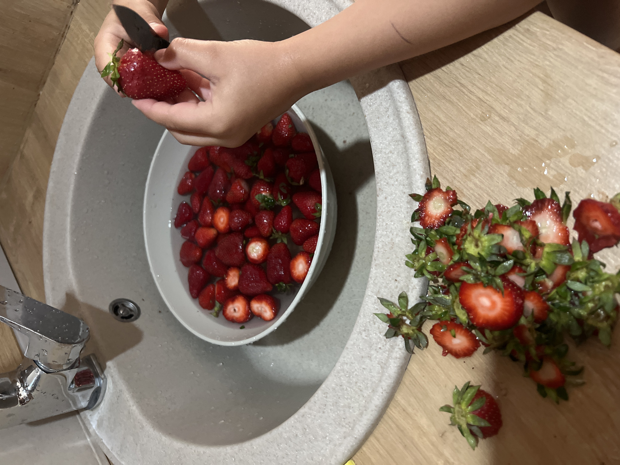 A person's hands are depicted actively hulling strawberries in a kitchen setting, likely in Budva, Montenegro. One hand holds a large, ripe red strawberry while the other uses a small, dark paring knife to remove the green leafy top and stem. A white bowl filled with numerous red strawberries, some already hulled and soaking in water, rests within a light-colored kitchen sink. A chrome faucet is visible on the left side of the sink, with water droplets on its surface and the surrounding sink basin, indicating recent or ongoing washing. On the wooden countertop to the right, a pile of discarded green strawberry tops, many still attached to small pieces of red fruit, is accumulating. Additional water droplets are scattered across the wooden surface. The scene captures an indoor activity, possibly food preparation, under ambient or artificial lighting. No specific time of day or emotions are discernible, and no text is visible.