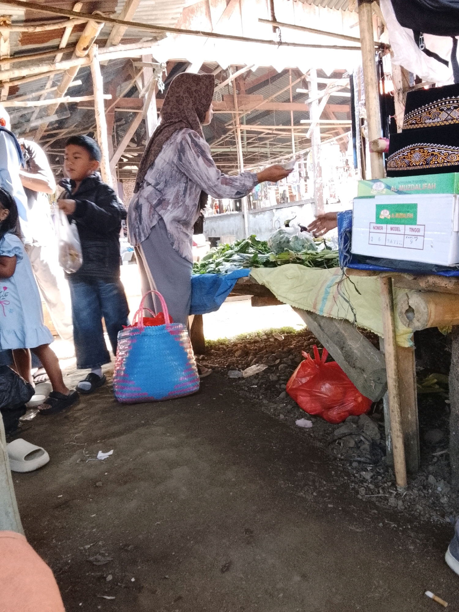 In an outdoor market stall in Sukarame, Indonesia, a woman wearing a patterned headscarf and gray top is shown at the counter, holding a white object in her right hand and extending her left hand as if completing a transaction. A young boy in a black jacket and blue pants stands to her left. A young girl in a light blue dress is also present, and a blue and pink woven basket sits on the ground in front of the woman. The market stall features a wooden structure with a corrugated metal roof and is filled with produce. A red plastic bag rests on the ground near the stall.