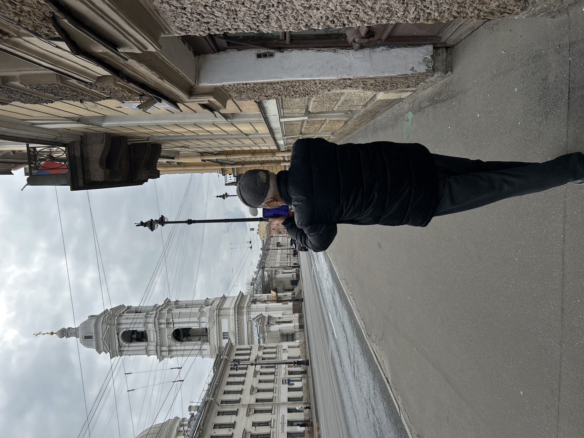 The image, correctly oriented, depicts a city street scene in Saint Petersburg, Russia, on an overcast day. A person, seen from behind, is walking along a wet asphalt sidewalk on the right side of the frame. The individual is a man wearing a dark puffer jacket, dark trousers, and a baseball cap, holding a smartphone horizontally in front of him, possibly capturing a photo or video of the surroundings.
To the left, an imposing white Baroque-style bell tower with a green dome and a golden spire topped with a cross dominates the background, strongly resembling the Saint Nicholas Naval Cathedral (Nikolsky Cathedral). The street is lined with multi-story buildings featuring classical architecture, ornate cornices, and numerous windows. Some of the building facades closer to the viewer show signs of wear, including rough textures and peeling paint.
Numerous overhead wires crisscross the grey sky, indicating urban infrastructure likely for public transport like trams or trolleybuses. Street lamps are visible along the sidewalk. The wet pavement suggests recent rain. On a building further down the street, a sign with Cyrillic text is visible, with the word "ЦЕНТР" clearly legible. The overall atmosphere is subdued under the cloudy sky.
