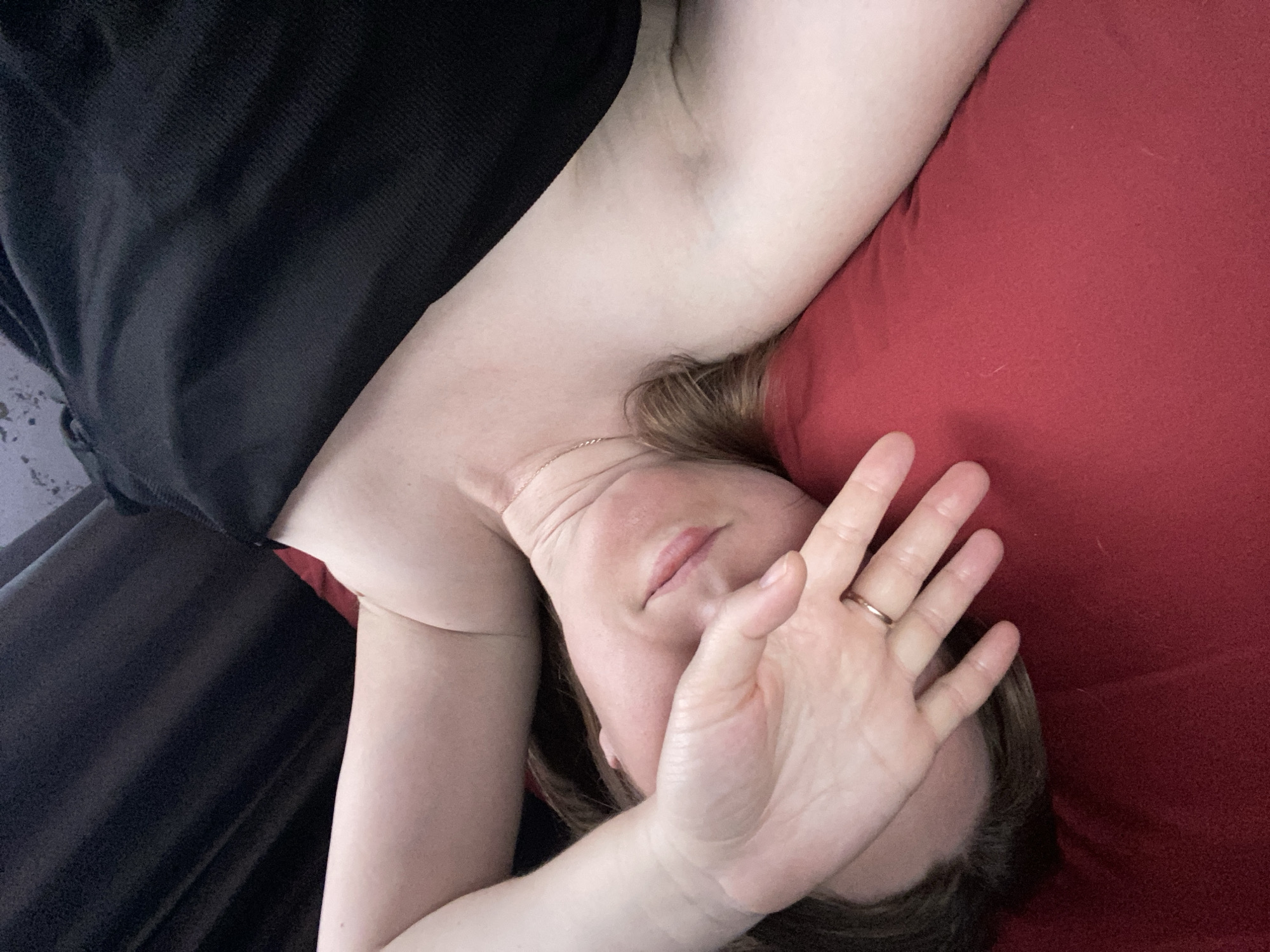 An overhead, slightly tilted shot captures a person lying down, likely indoors in Noviy, Russia. The individual, appearing to be an adult, is positioned with their head turned towards the right side of the frame, partially obscured by their raised left hand which covers most of their face. The fingers are slightly spread, and a simple gold-colored ring is visible on the ring finger.
The person has light skin and shoulder-length, light brown or auburn hair that falls onto a red fabric surface, which appears to be a pillow or bedding. A delicate gold-colored necklace is visible around their neck. The top portion of their body, including an arm and shoulder, is visible. They are clad in what appears to be dark, possibly black, fabric or clothing that covers the upper left side of the frame. The lighting is soft and even, suggesting an indoor setting without harsh direct light.
No specific activity beyond lying down is clearly depicted, though the hand over the face might suggest shyness, privacy, or blocking light. There is no visible text in the image. The scene conveys a sense of quiet intimacy or repose within an indoor environment.