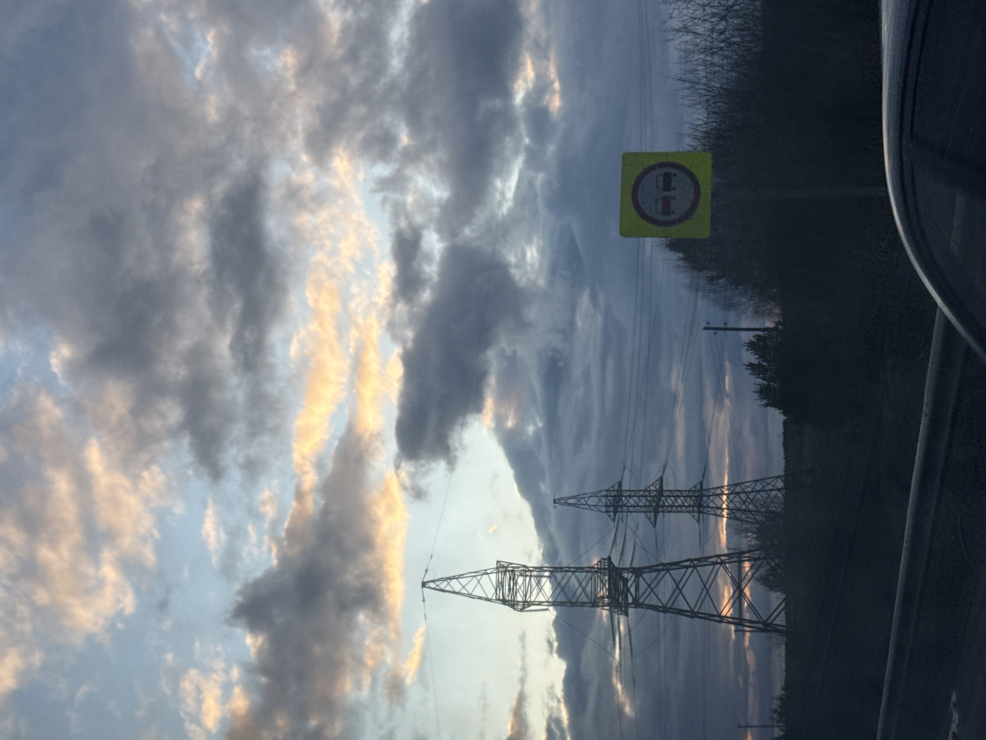 The image, taken in Гаревая, Russia, captures an outdoor landscape scene, likely from a vehicle during sunrise or sunset. The sky dominates the frame, filled with dramatic, layered clouds. Patches of the clouds are illuminated with warm, golden-orange light, contrasting with extensive areas of darker gray and blue.
In the lower portion of the image, two large electricity pylons (power line towers) stand against the luminous sky, their intricate metal structures appearing as dark silhouettes. Power lines extend between them and across the scene. To the right, a square yellow road sign is visible. It features a red circular border enclosing a graphic of two dark vehicles (likely cars or trucks) positioned side-by-side, indicating a "no overtaking" or "no passing" zone.
Behind the sign and along the right side of the frame, a dense line of dark trees or bushes forms a natural barrier. The extreme right edge of the image reveals a curved, dark surface, suggesting it is part of the vehicle from which the photograph was captured. The overall impression is of a vast, open sky over a rural or semi-rural landscape during the twilight hours.