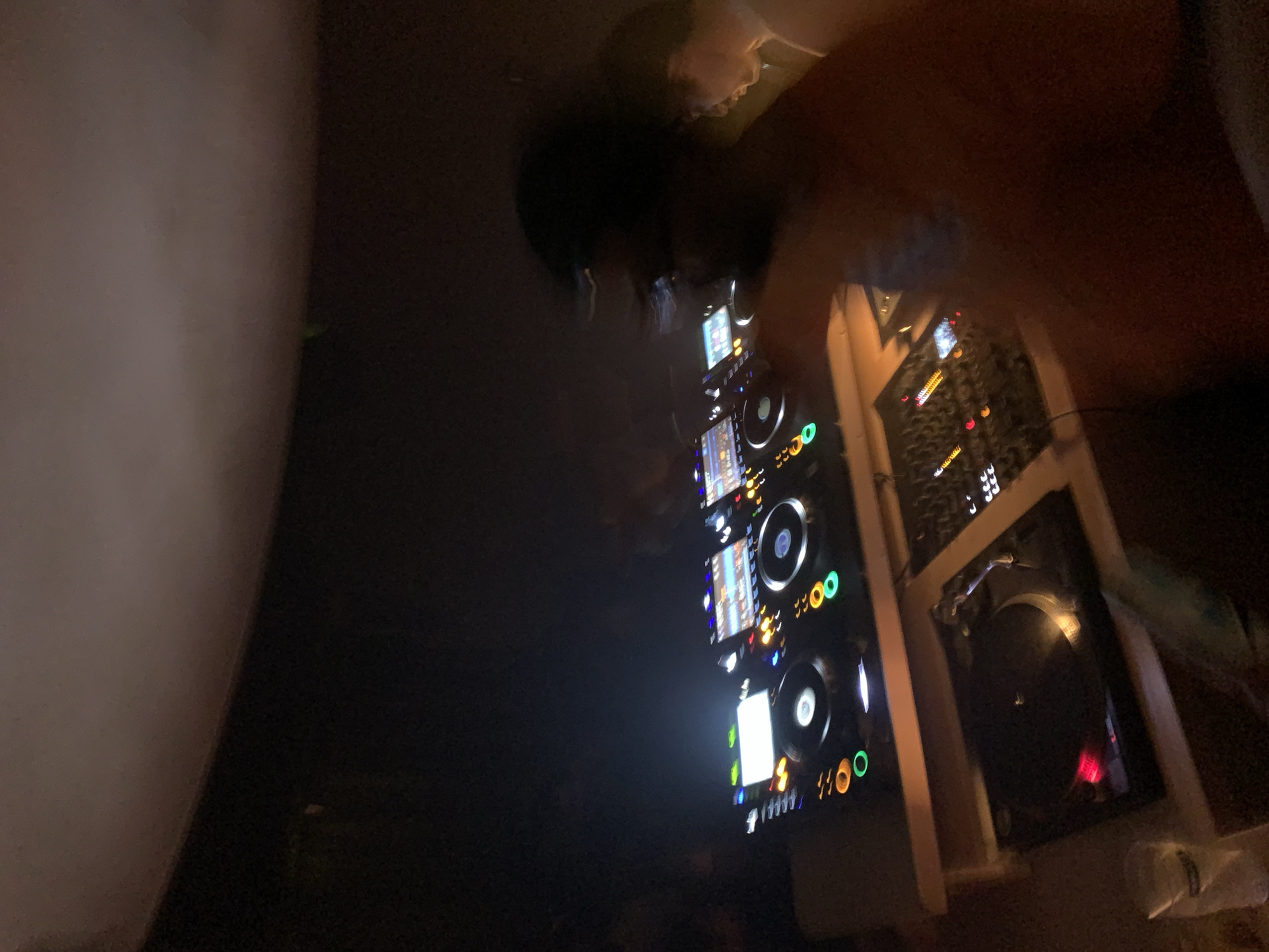 A low-angle shot in a dimly lit space, likely a club or event venue in Tel Aviv, Occupied Palestine, features a DJ setup. The setup comprises what appear to be three DJ CD decks, a mixer, and a turntable. The decks are vertically stacked and illuminated with colorful lights. A person is partially visible in the top center, possibly the DJ in action. The surrounding environment appears dark and undefined.