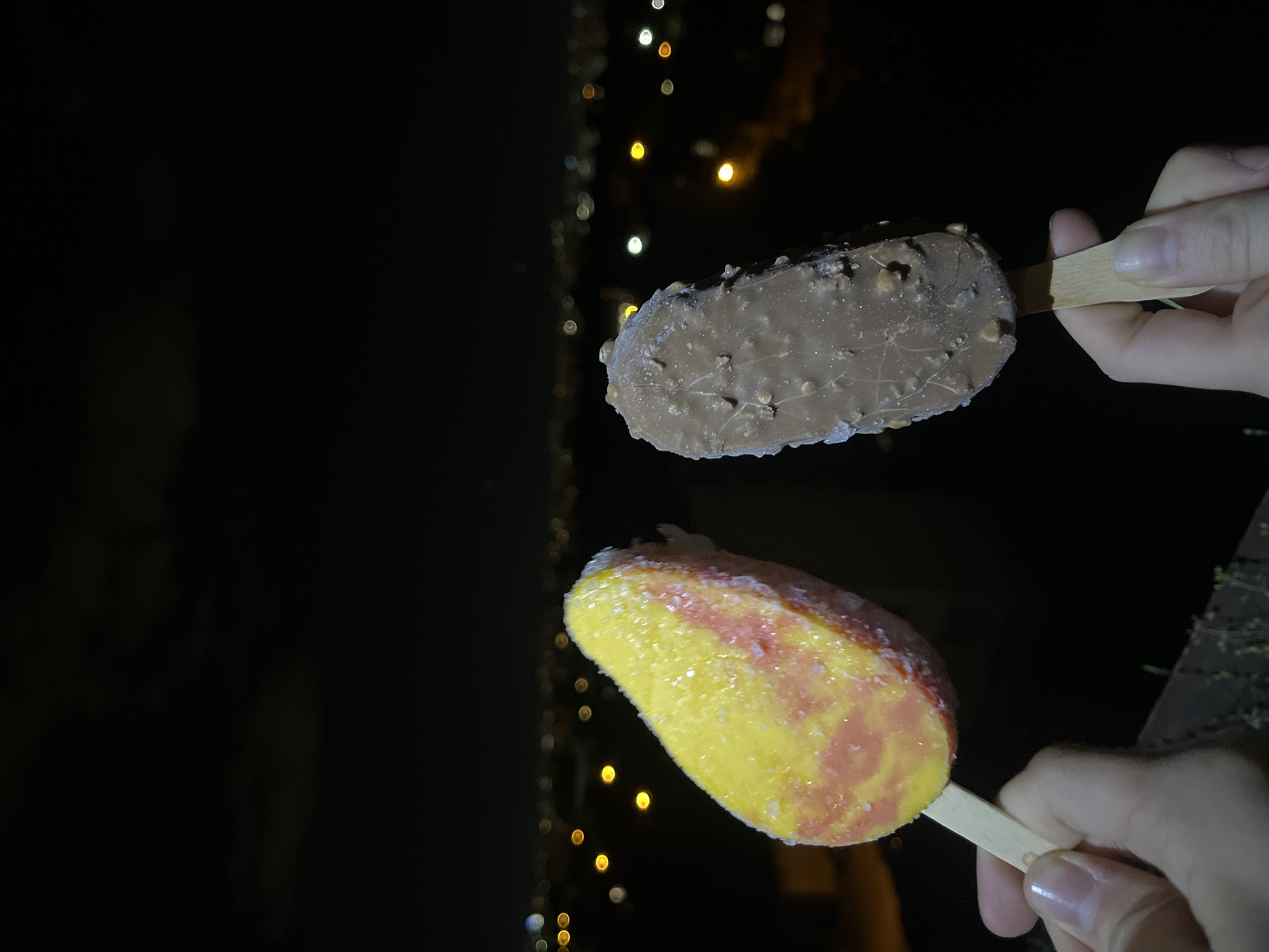 The image shows two hands each holding an ice cream bar on a wooden stick. The upper ice cream bar is brown, likely chocolate, and appears to have nuts embedded in the coating. The lower ice cream bar is yellow with swirls of red or orange, suggesting a fruit flavor like mango or peach. The background is dark with blurred lights, which suggests an outdoor setting at night. The lights in the background are likely city lights and could be from Sofia, Bulgaria, but without further context, it is difficult to confirm this location definitively.