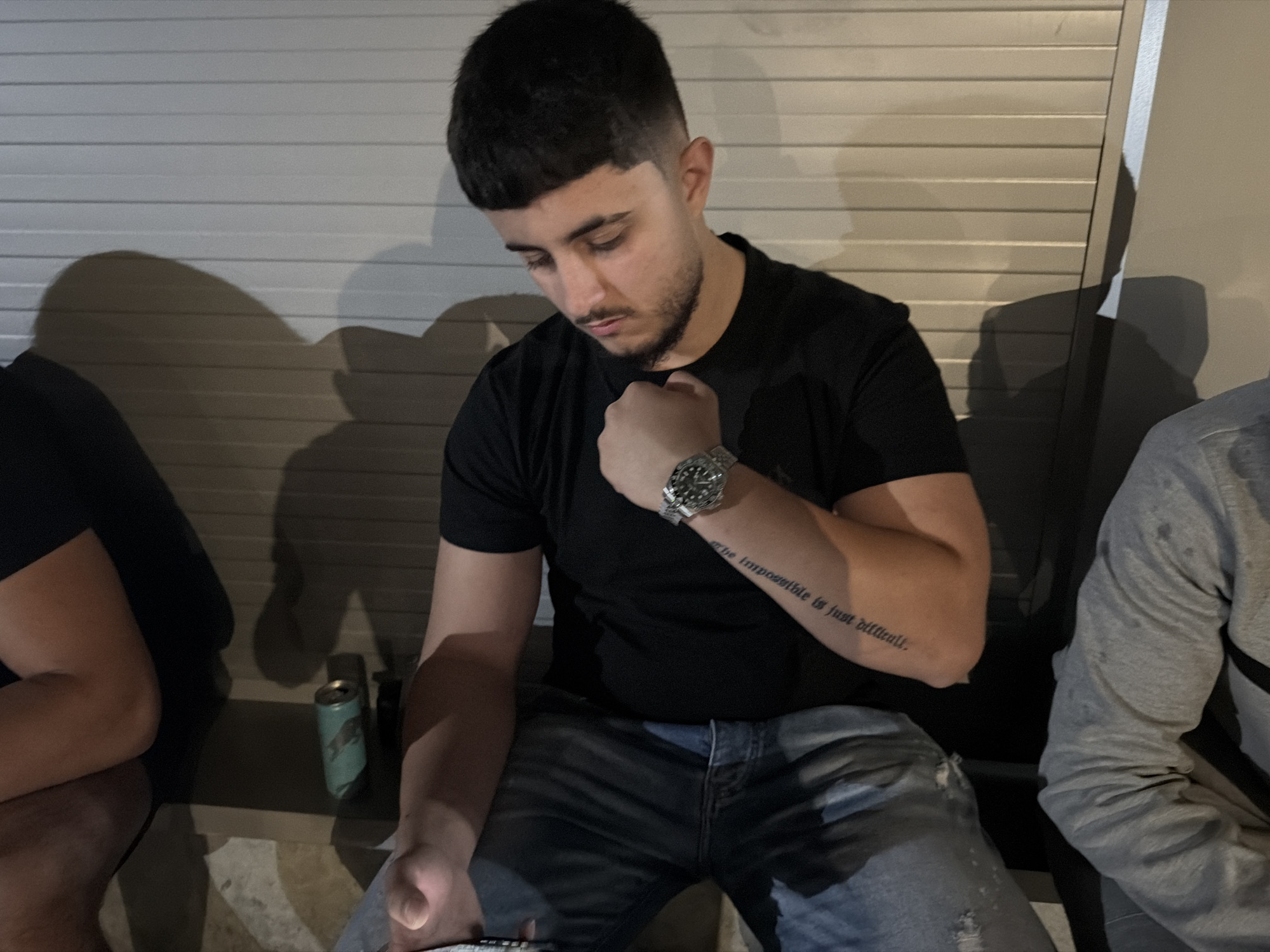 The image captures a young man, likely in his late teens or early twenties, seated and looking down with a focused and serious expression. He has short dark hair styled with a bowl cut and faded sides, and sports a beard and mustache. He is wearing a black short-sleeved t-shirt and blue ripped jeans. A silver watch is visible on his left wrist, and his left hand is clenched against his chest. On his left forearm, a tattoo reads: "The impossible is just difficult."
Partial views of other individuals are visible on either side of the main subject, with arms and shoulders suggesting a casual gathering. A light blue/green aluminum can is placed on the ground between the main subject and the person to his left. The background features a light-colored, horizontally slatted wall, possibly a garage door or a shutter. Strong shadows are cast on this wall, indicating artificial lighting and suggesting the scene takes place at night or in a dimly lit indoor or sheltered outdoor setting. The overall atmosphere is informal.