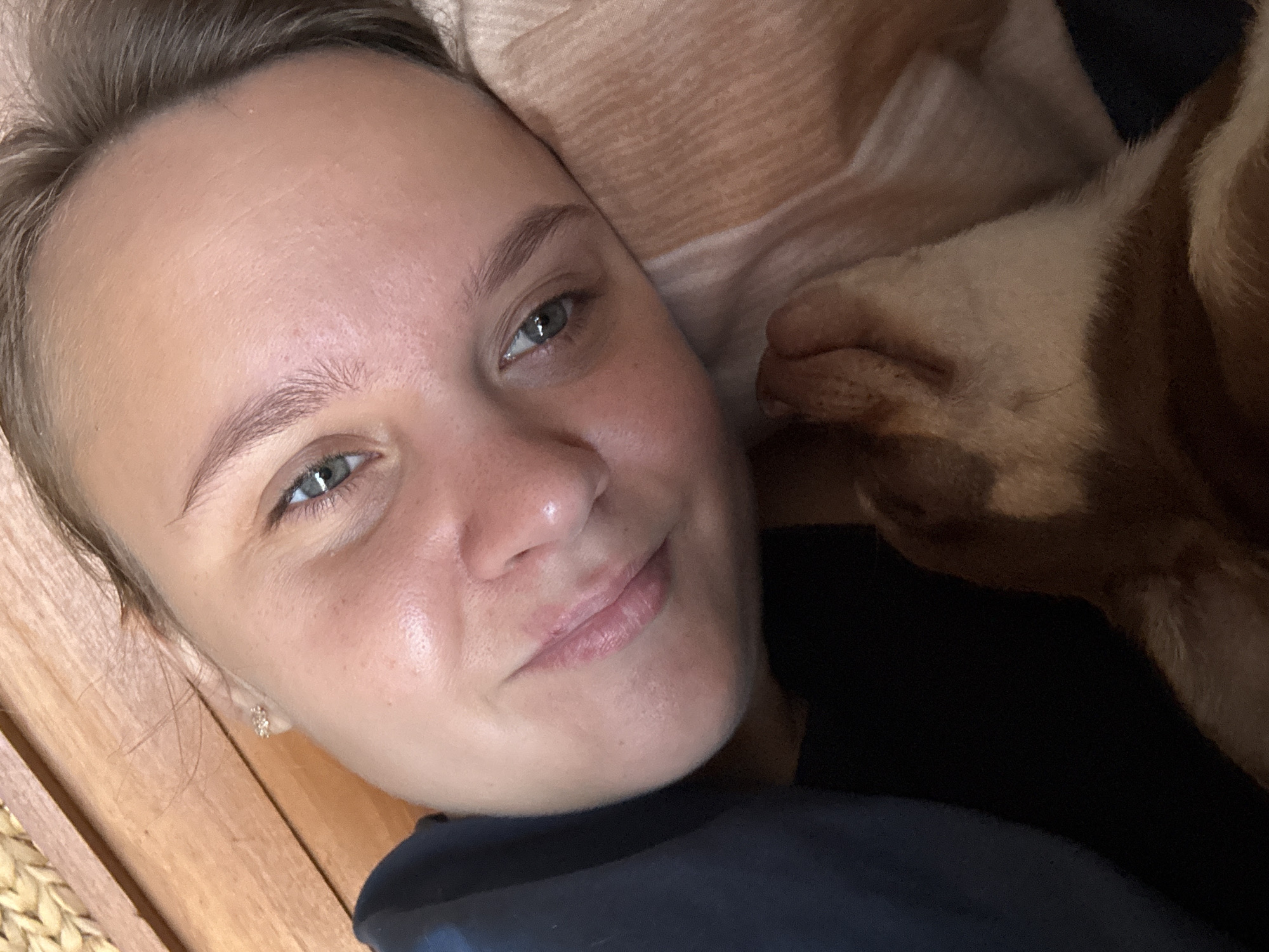The image shows a close-up, low-angle selfie of a young woman lying down, with her head resting on a pillow and her dog nuzzling against her face. The woman is looking up at the camera with a slight smile. Her left ear is visible, adorned with a small, sparkly earring. Her hair is light brown and pulled back from her face. The dog, a light brown and white breed, is positioned in the lower right quadrant of the frame, with its nose touching the woman's cheek. The background consists of a wooden surface on the left and a patterned blanket or pillow on the right. The lighting appears to be natural, casting a soft glow on the subjects. The scene suggests a relaxed and affectionate moment between the woman and her pet.