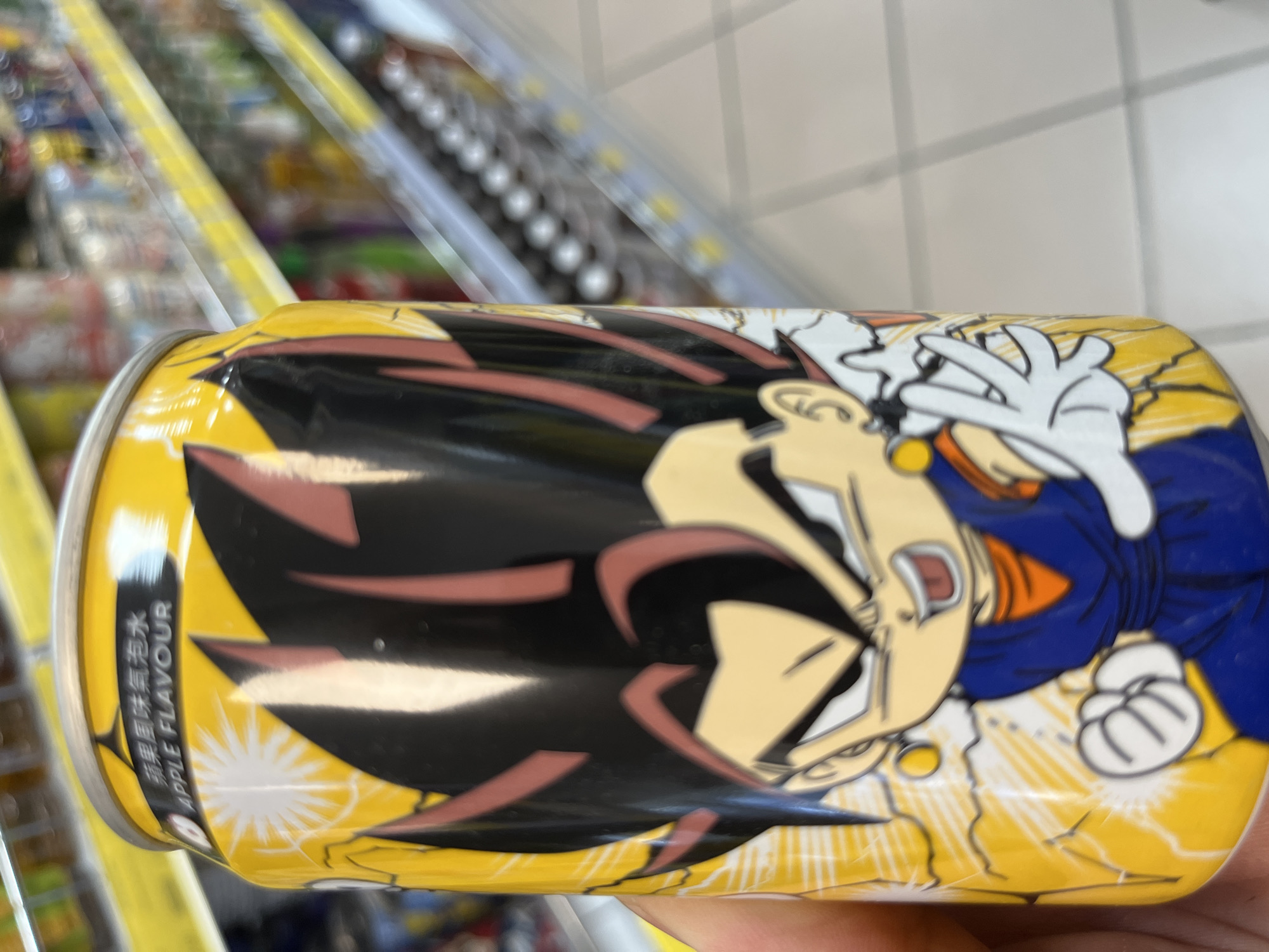 A hand holds a yellow soda can featuring an anime character. The can has Chinese characters and the words "APPLE FLAVOUR" in English. The background shows blurred shelves stocked with beverages in a retail environment, indicative of a store. The image was taken in Castiglione delle Stiviere, Italy.