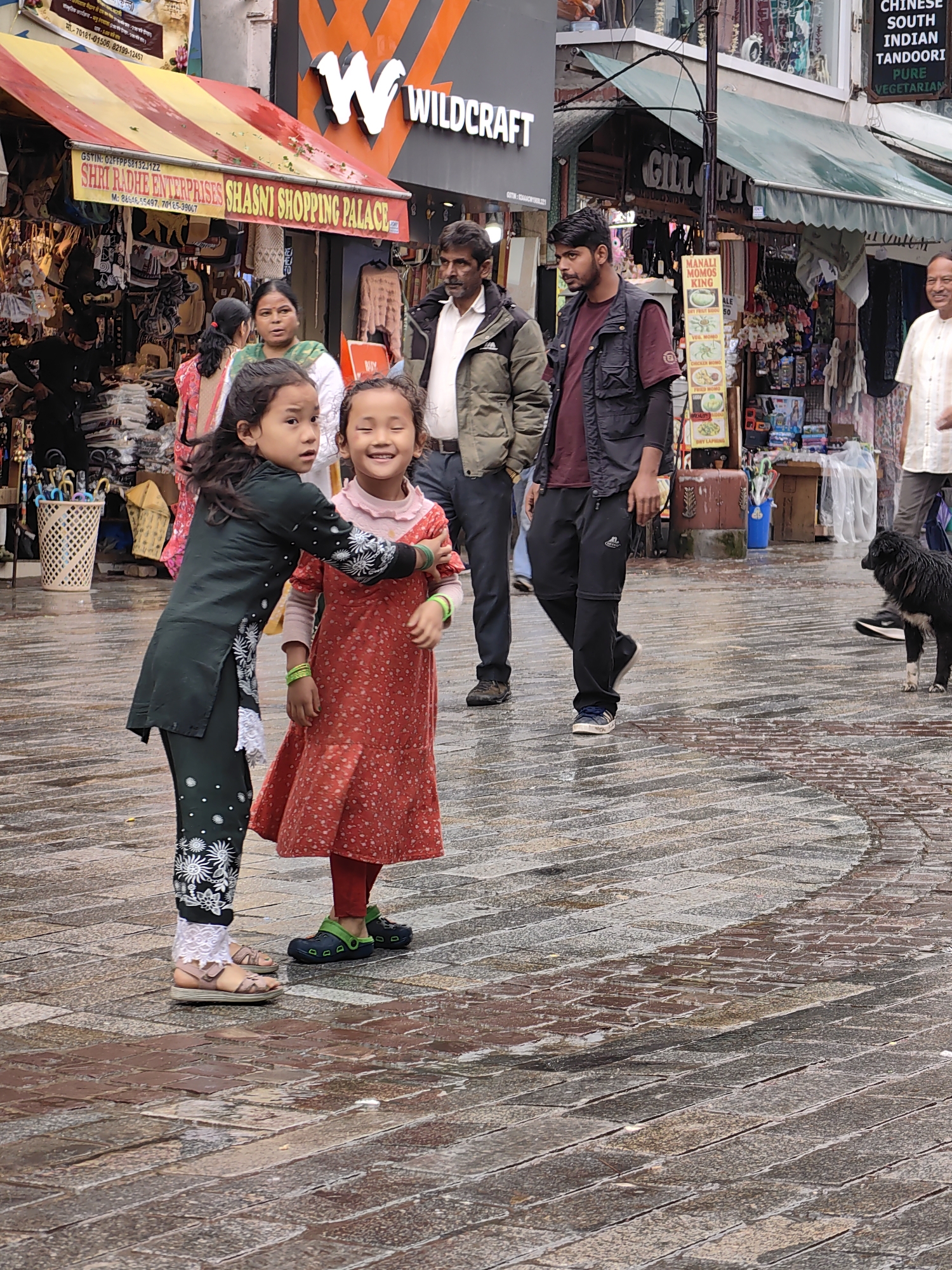 This image captures a lively street scene in Manali, India, likely on The Mall Road, given the presence of shops and pedestrians. Two young girls are the central focus. The girl on the left, wearing a dark green traditional Indian outfit adorned with white floral patterns and lace trim, has her arm around the girl on the right. The girl on the right is wearing a vibrant red, polka-dotted dress with a pink collar and red leggings. She is smiling broadly, with her eyes closed, suggesting she is laughing or reacting to something playful.
Several adults are also visible in the background. To the left, a woman in a light green and white outfit stands with her back to the camera. Next to her, a man in a light brown jacket and dark pants appears to be looking towards the children. To the right of the children, a man in a black vest over a maroon shirt and black pants is walking. Further to the right, another man in a light-colored shirt is walking away from the camera.
The environment is a wet, paved street, indicating it may have recently rained. The cobblestone-like pavement is reflective with puddles, adding to the atmosphere of a bustling marketplace. Shops are visible on either side of the street, with awnings and signs indicating their presence, including one for "SHRRI RADHE ENTERPRISES SHASNI SHOPPING PALACE." In the background, a black dog with white markings is walking on the wet pavement. The overall impression is one of everyday life unfolding in a busy tourist area.