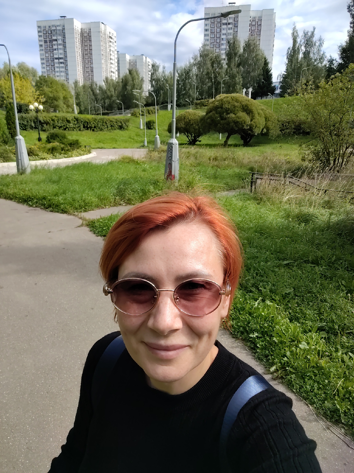 In the city of Zelenograd, Russia, a woman with short, reddish-orange hair and rose-tinted sunglasses smiles at the camera in a selfie. She wears a black, long-sleeved top and a blue backpack strap is visible over her right shoulder. She stands on a paved path, with a lawn and trees to her right. In the background, tall residential buildings and streetlights are visible under a partly cloudy sky. The scene appears to be a park or public area within a residential district.